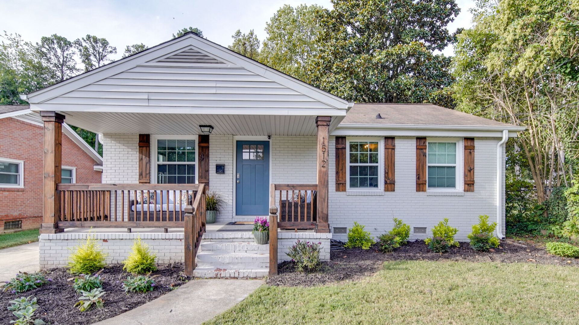 Single-story white brick house with a covered front porch, blue front door, wooden railings and shutters, and a manicured lawn with small shrubs along the foundation.