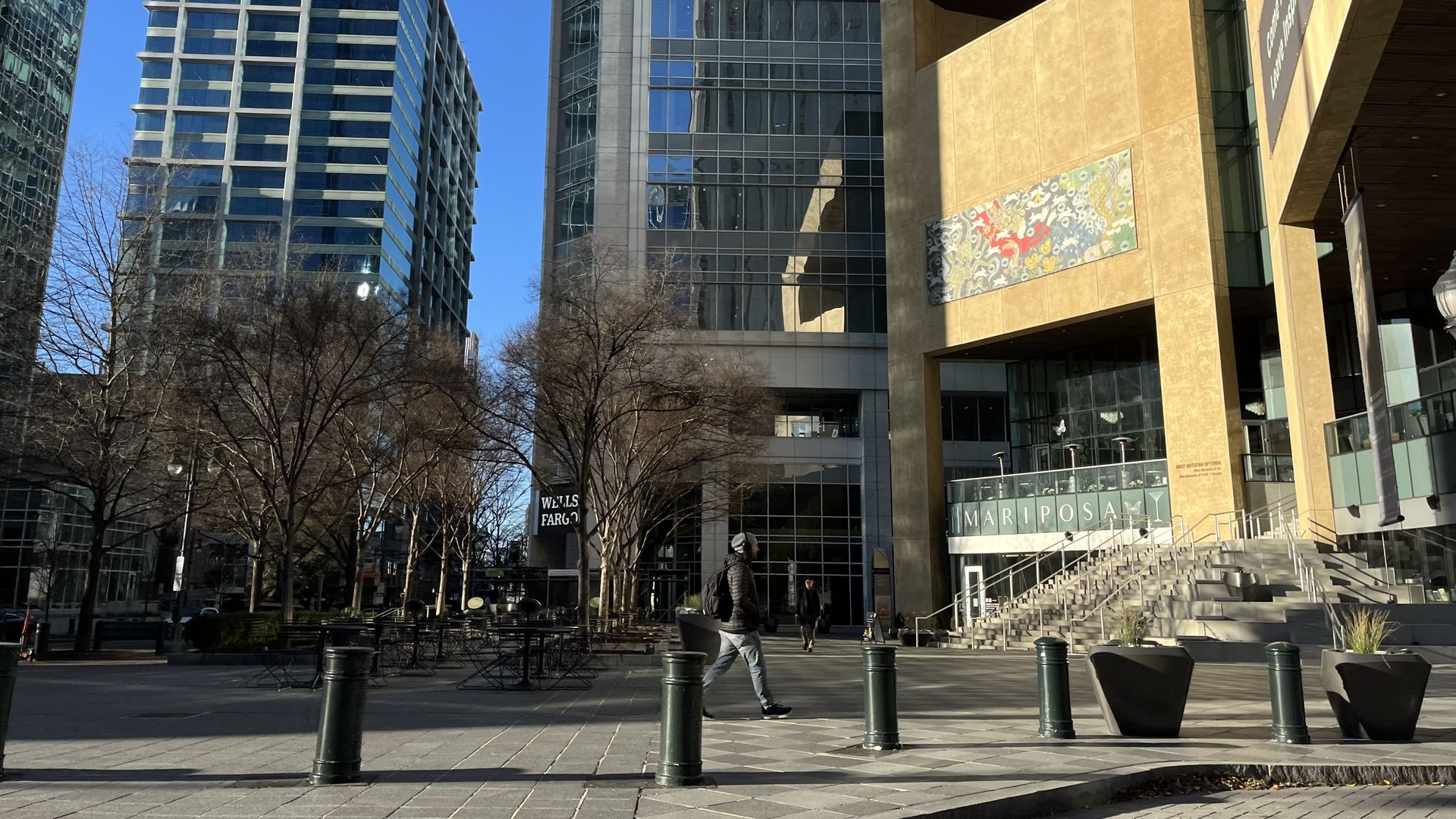 Urban plaza with leafless trees, outdoor tables, a person walking, tall modern glass buildings, and the Mariposa building with a gold facade and artwork panel under clear blue sky.