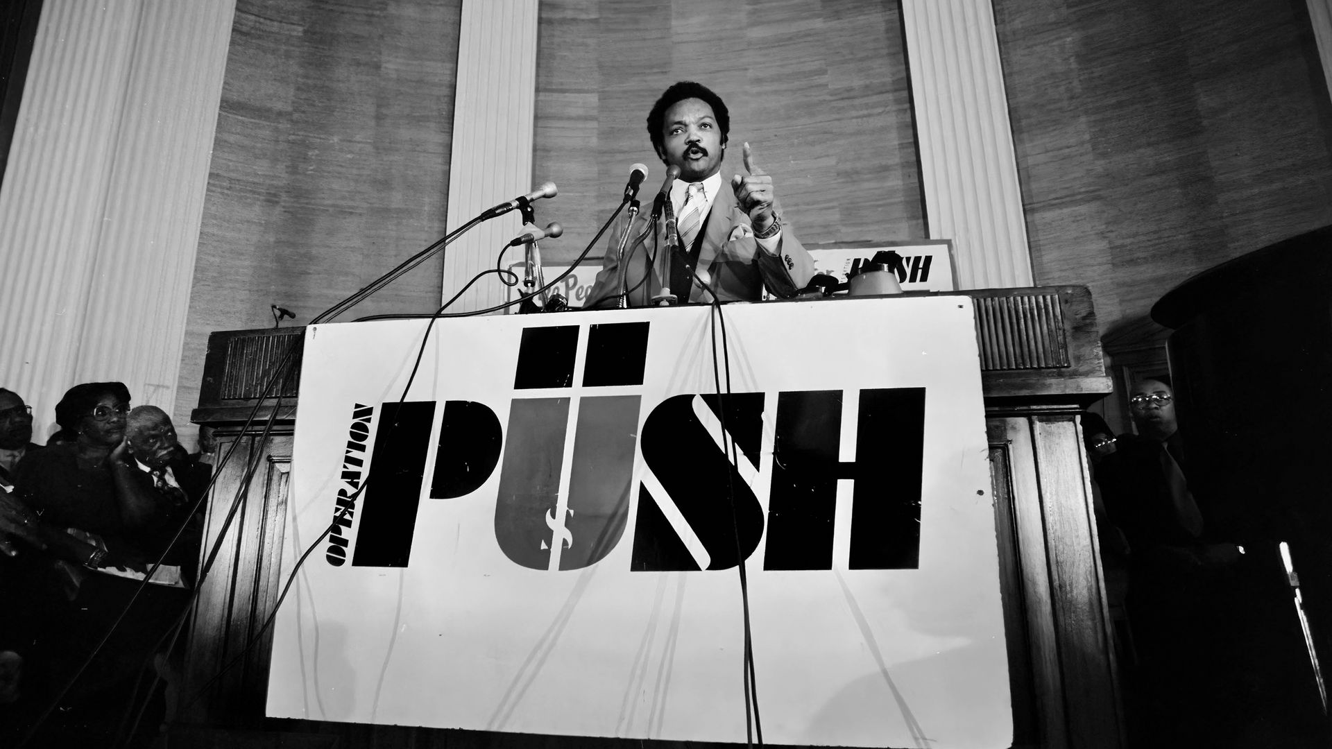Black and white photo of a man in a suit speaking passionately at a podium with microphones, behind a sign reading "Operation PUSH" in bold letters.