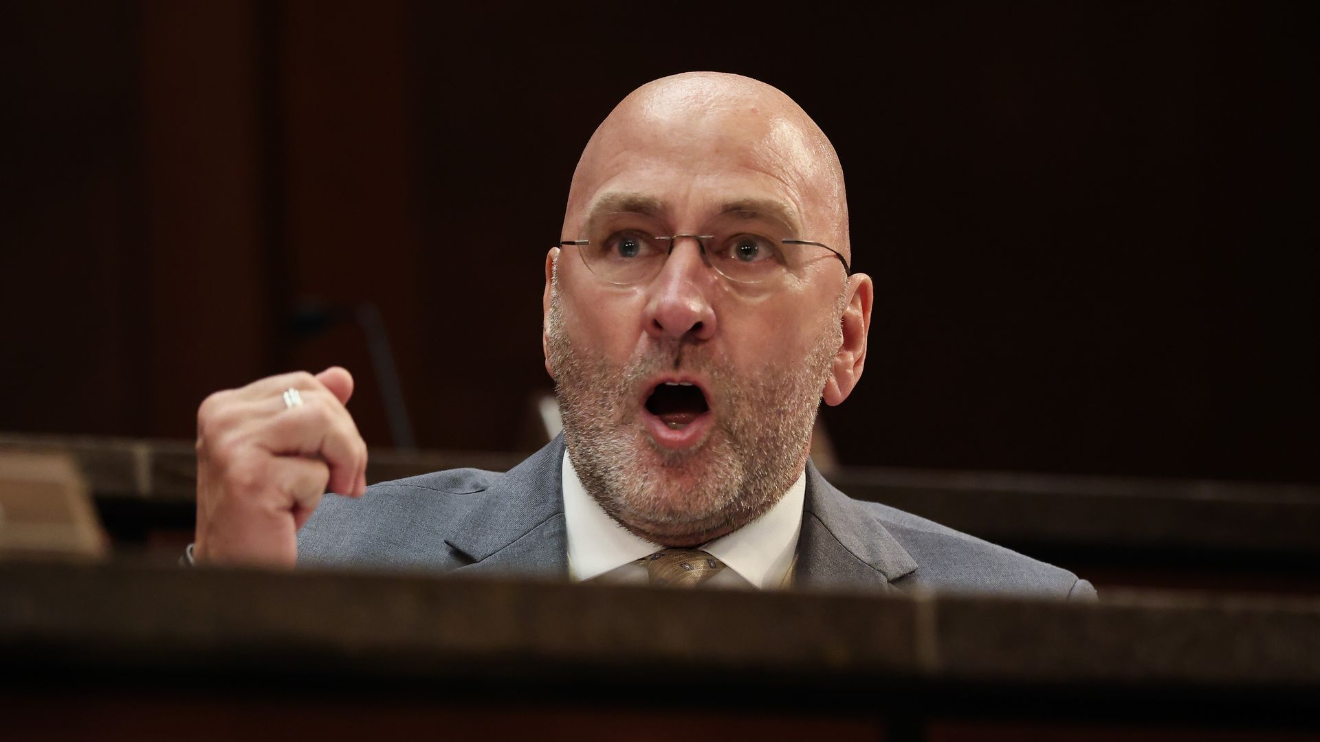 Bald man with glasses, beard, and a gray suit raises fist and speaks emphatically in a dark indoor setting, against a dark background with partial stone barrier visible.