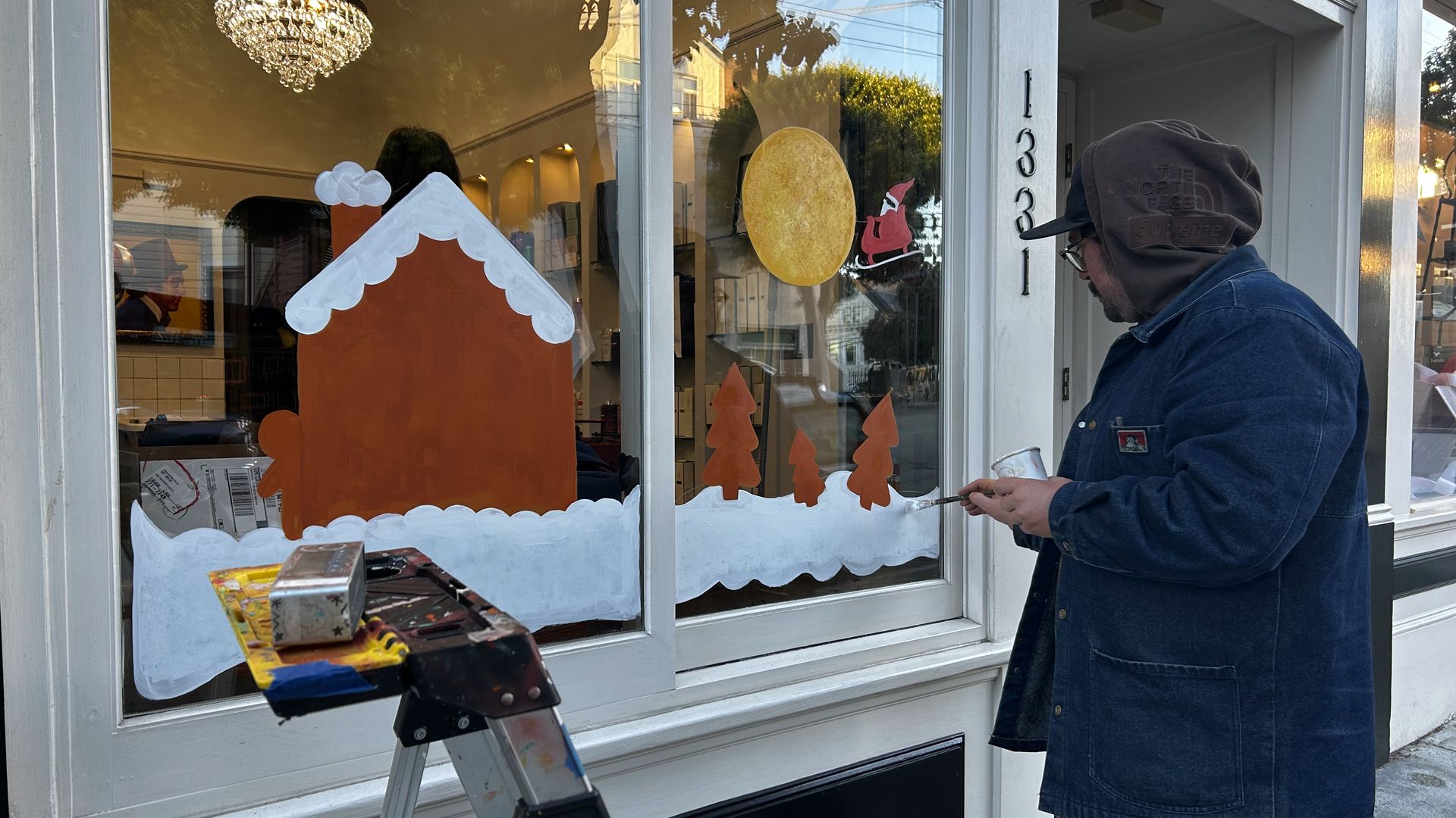 Person in denim jacket and brown hood painting Christmas-themed window art with brown house, white snow, orange trees, yellow moon, and Santa in red sleigh on building numbered 1331.