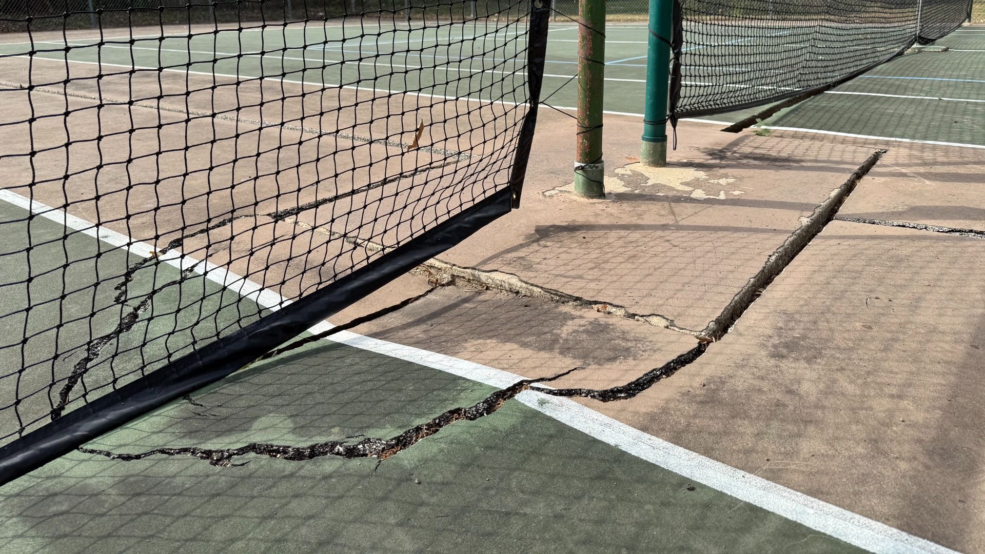 Close-up of a cracked and damaged tennis court surface with two separate nets, green and tan court colors, and visible large cracks near the net posts.