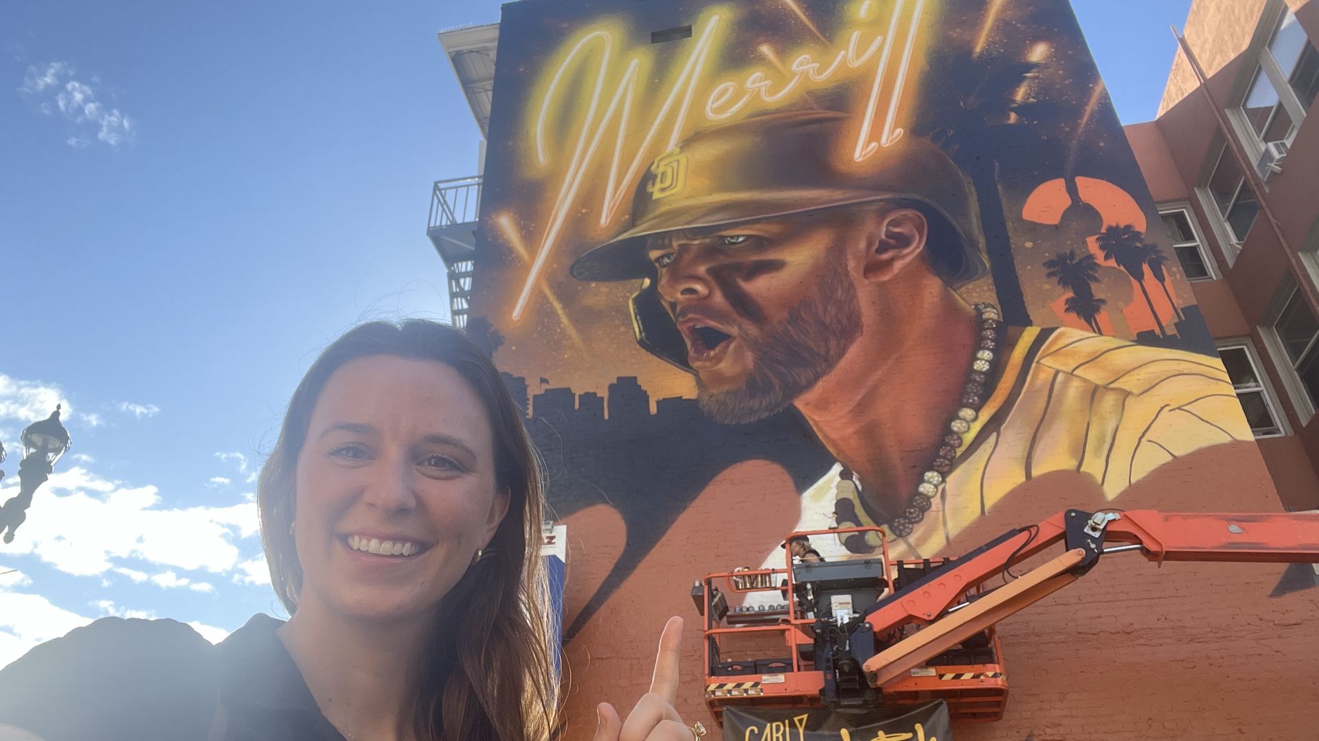 Smiling woman points to a large colorful mural of a baseball player wearing a brown Padres helmet, beaded necklace, and eye black on a building wall with the words "Merrill" in neon writing, under blue sky with light clouds.