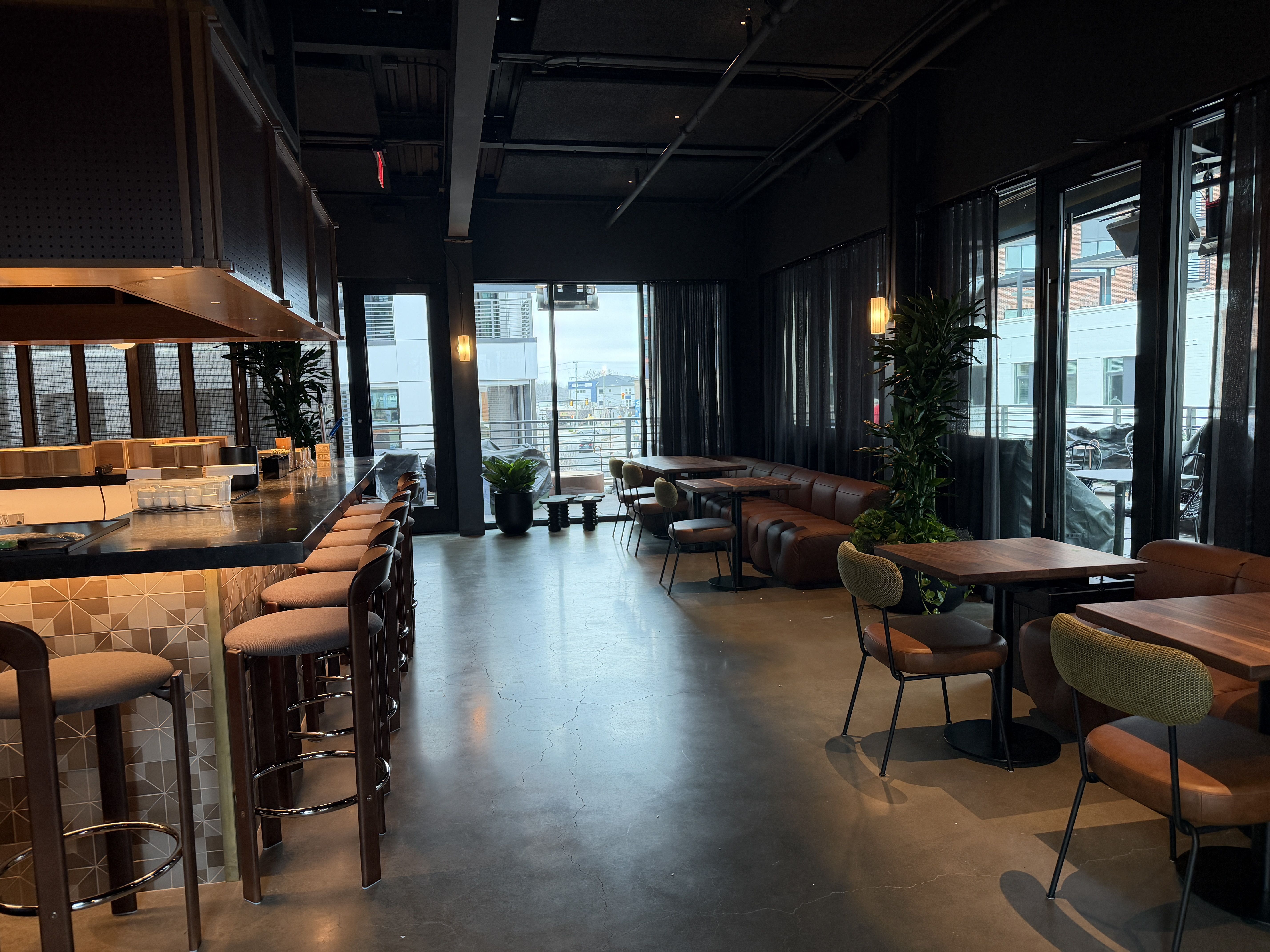Empty modern bar and lounge with brown leather stools and chairs, wooden tables, black ceiling, large windows with sheer black curtains, and potted green plants.