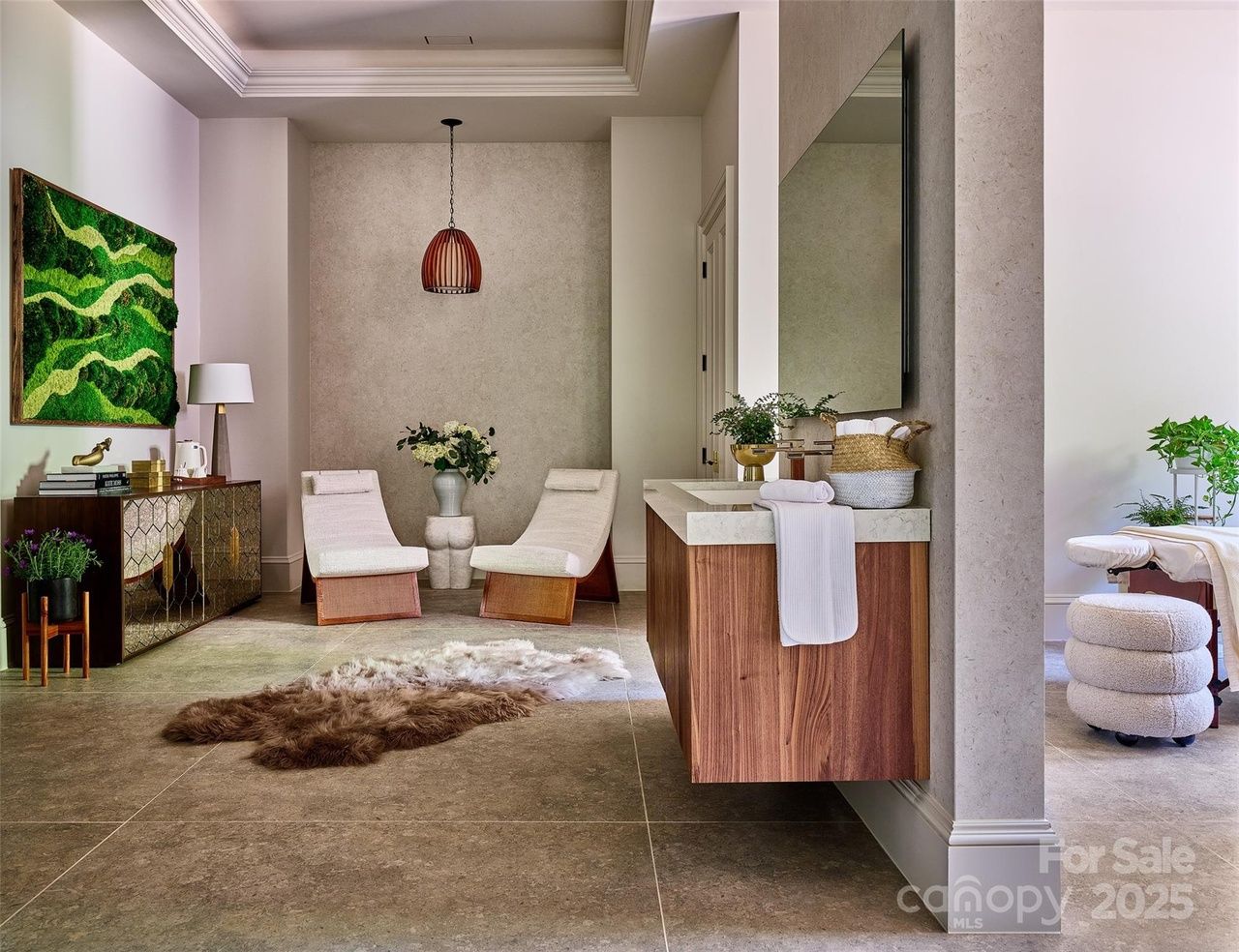 Modern living room with two white cushioned wooden chairs, a green moss wall art, a brown and white fur rug on tiled floor, and a wooden cabinet with a white lamp and books.