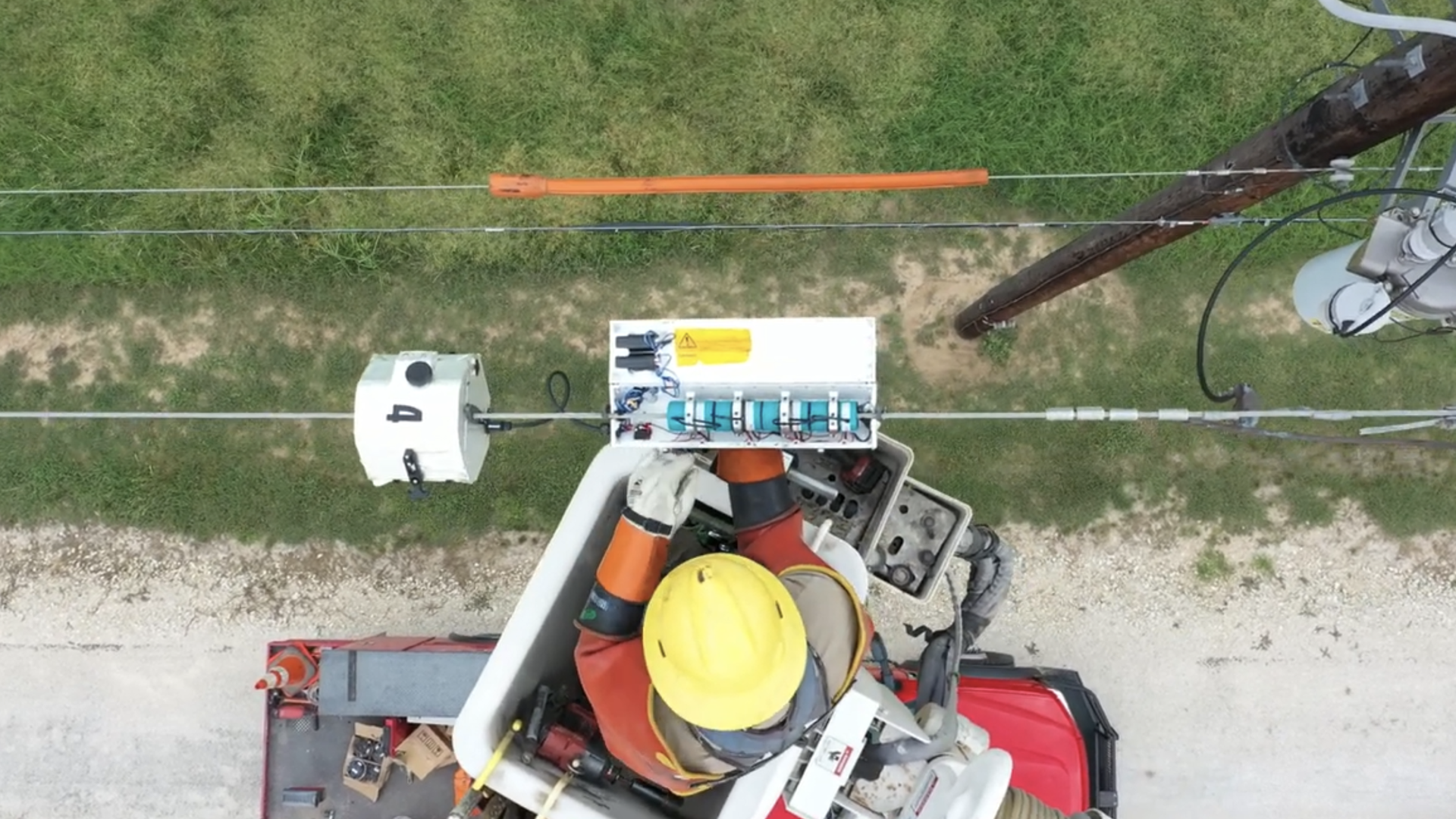 Aerial view from a bucket lift showing workers in yellow helmets and orange jackets on a utility pole, repairing power lines. An orange protective sleeve spans the line; equipment rests on grass.