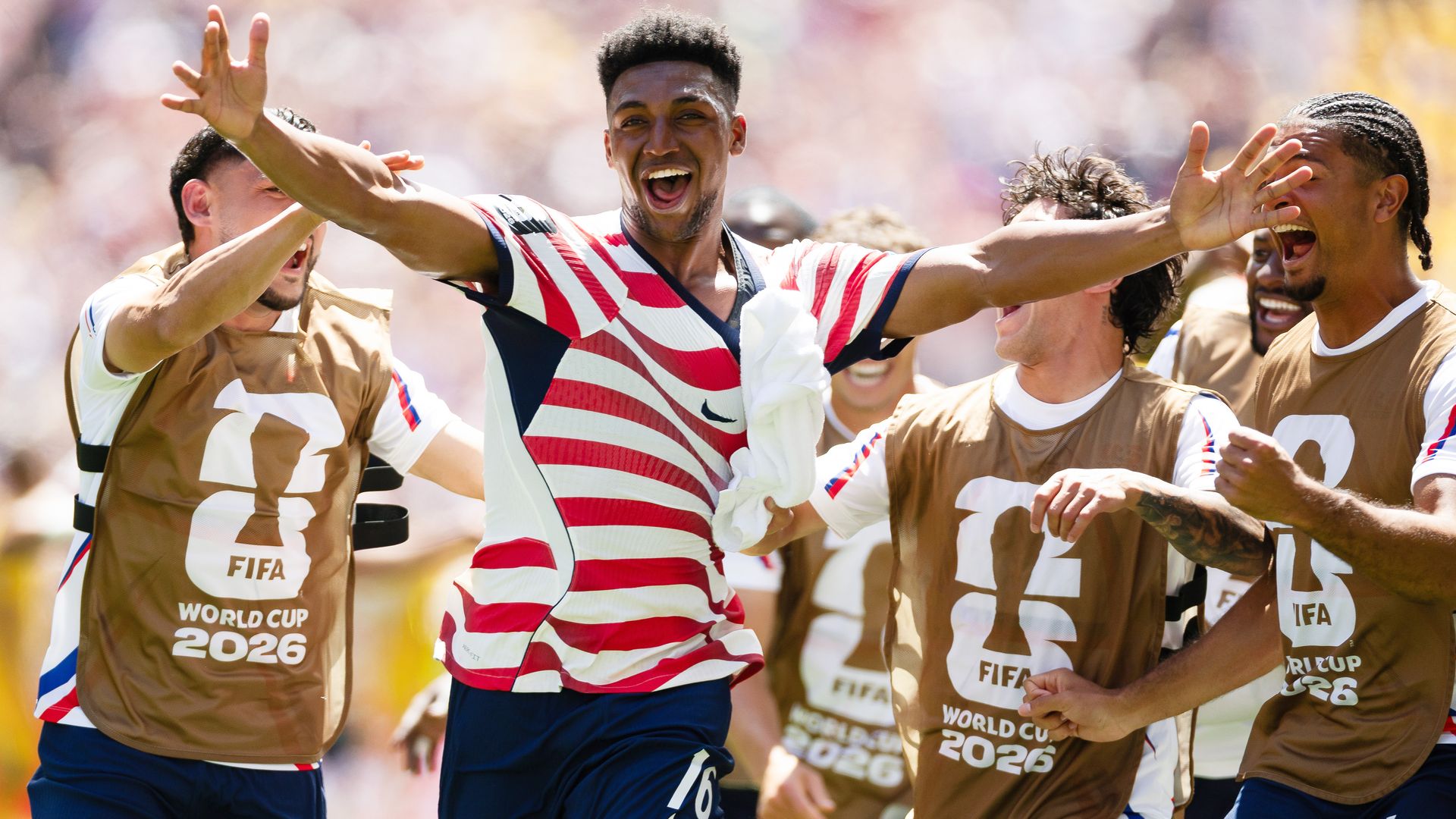 Alex Freeman with his arms outstretched, with his mouth open smiling surrounded by his teammates who are substitutes wearing the brown FIFA World Cup 2026 pinnys. They celebrated his goal against Australia.