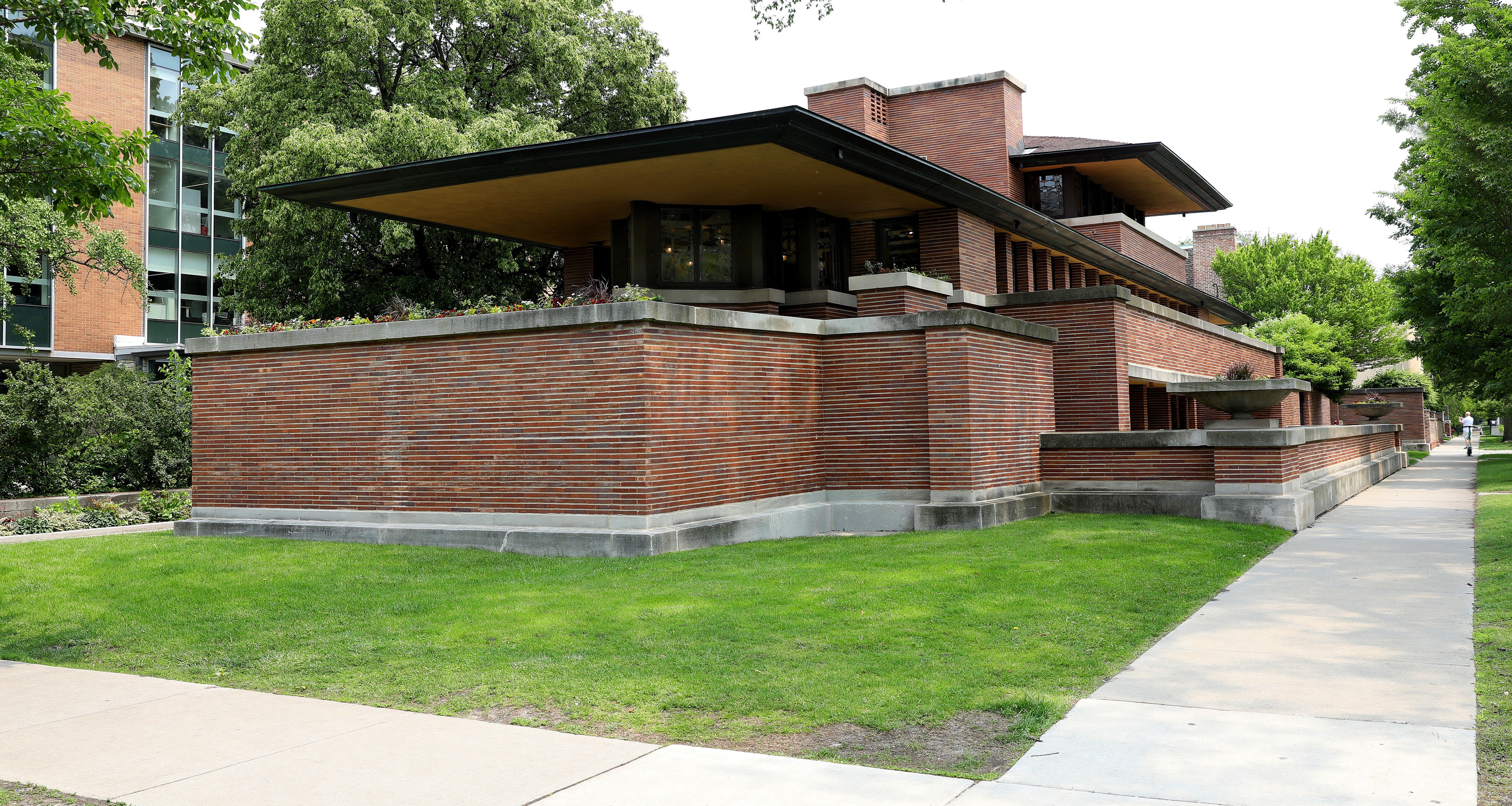 Frank Lloyd Wright's Robie House. Low, horizontal brick building with wide, overhanging eaves in a lush green setting next to a concrete sidewalk, surrounded by trees and shrubs.