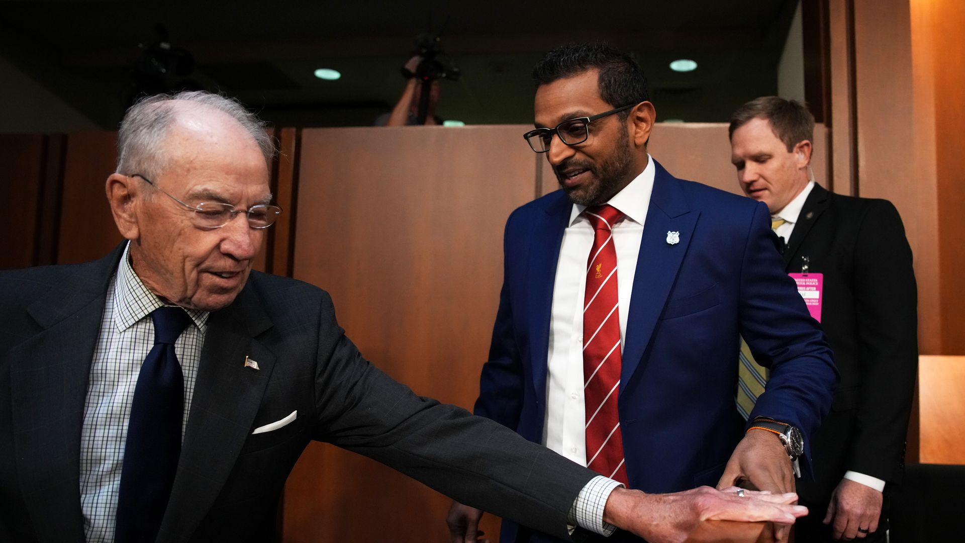 Grassley wearing a black suit jacket, blue and white shirt and blue tie. Patel wearing a blue suit jacket, white shirt and red and white striped tie. 