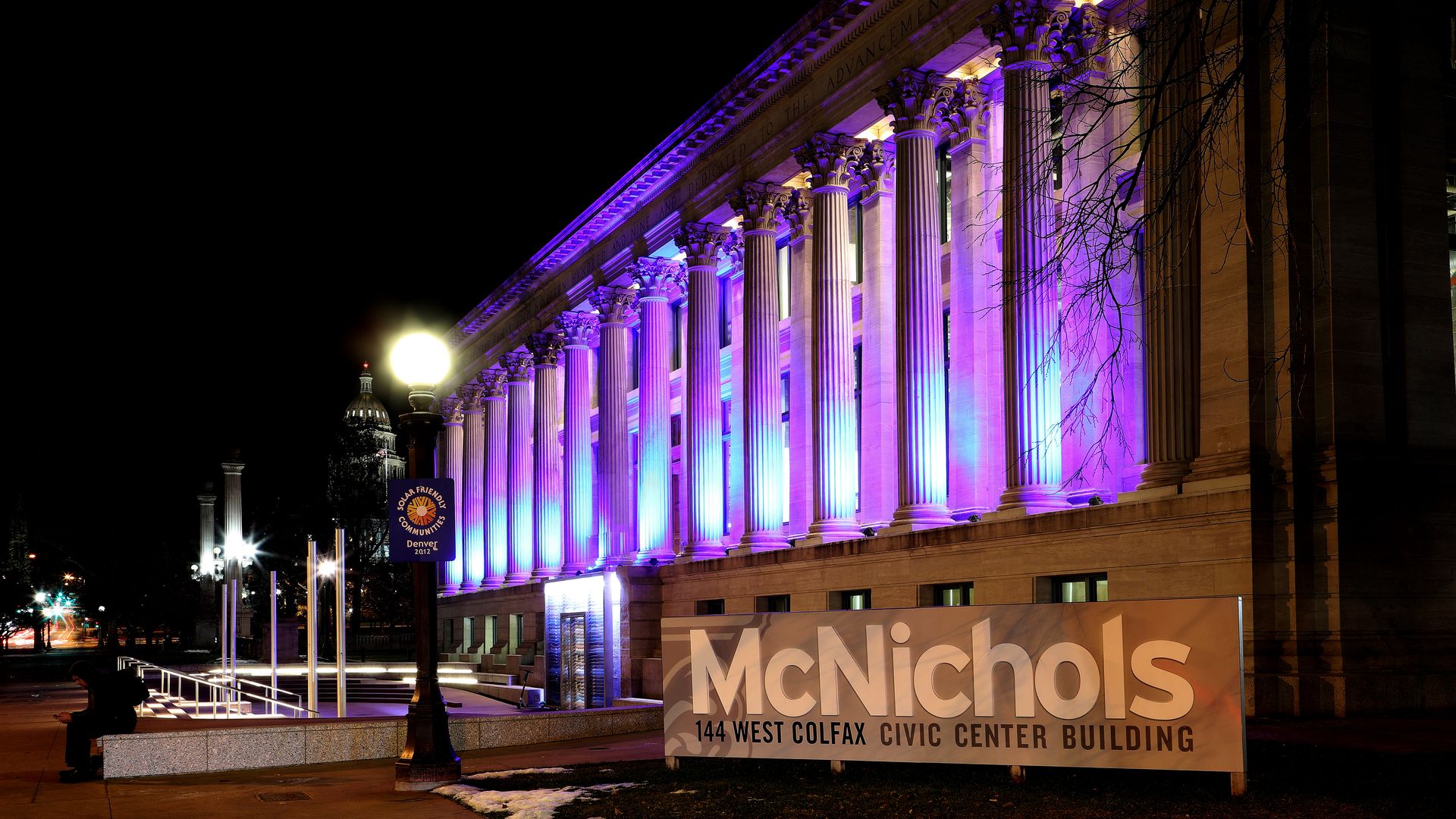 A neoclassical building lit in purple lighting is seen behind a sign reading MCNICHOLS 144 WEST COLFAX CIVIC CENTER BUILDING.
