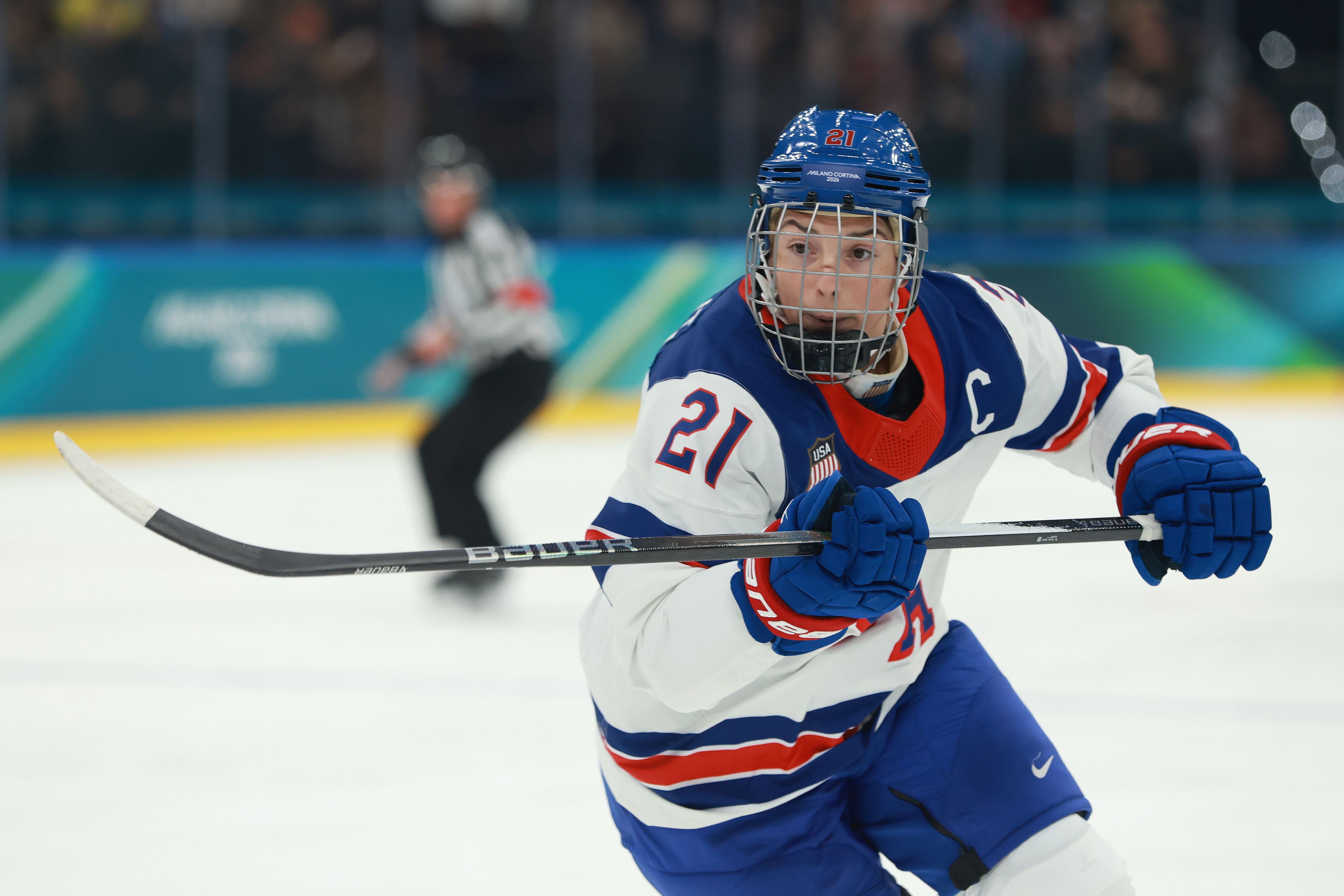 Hilary Knight leans forward with a hockey stick perpendicular to her body during a play on the ice, wearing a blue helmet with facemask. Her jersey is white red and blue and has the number 21 on it. 