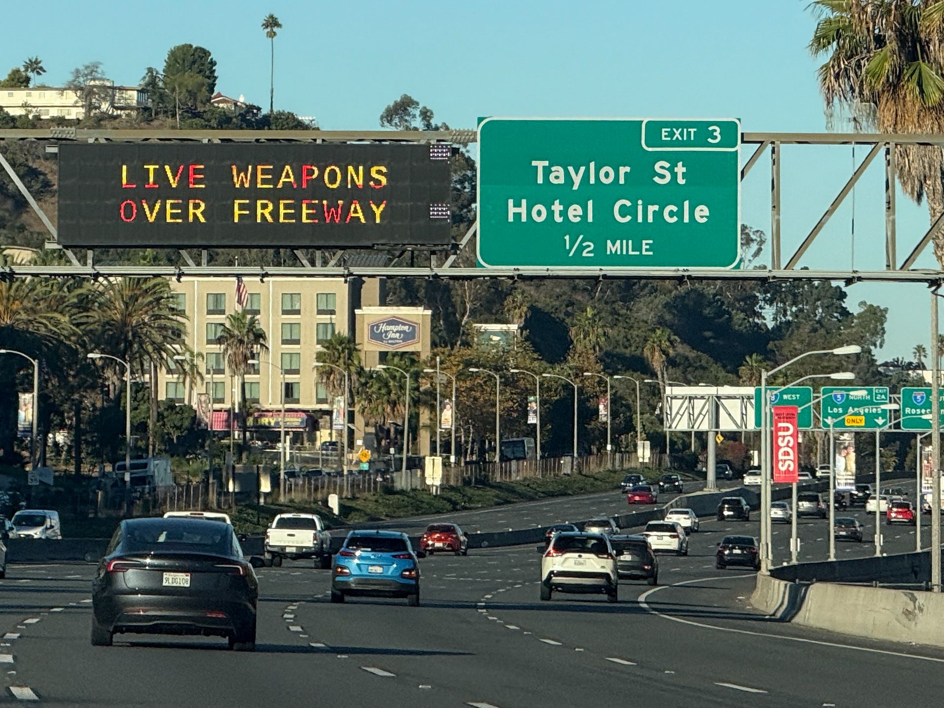 A traffic sign over Interstate 8 in San Diego says "LIVE WEAPONS OVER FREEWAY" as cars drive through approaching Interstate 5 towards Los Angeles.