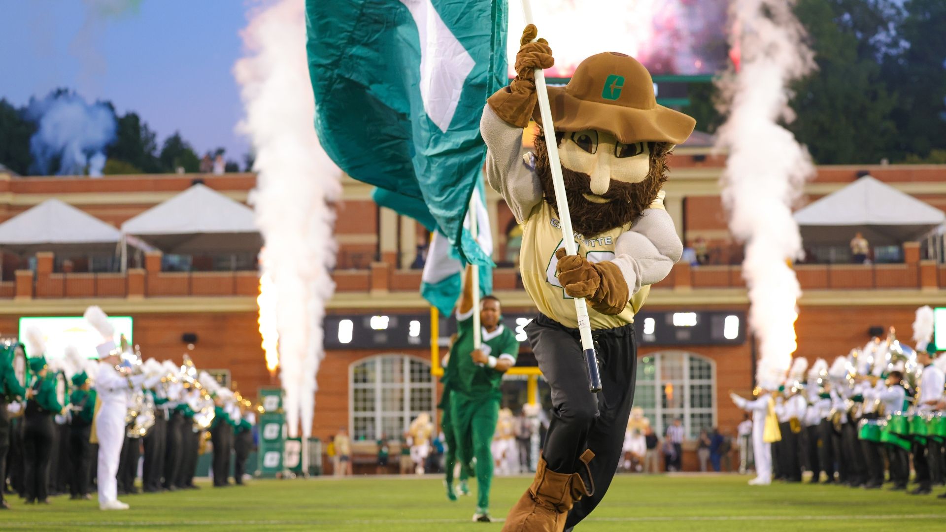 Mascot wearing tan hat and gloves with a flag walks on football field during a game, with band members in green and white uniforms and smoke effects in the background.