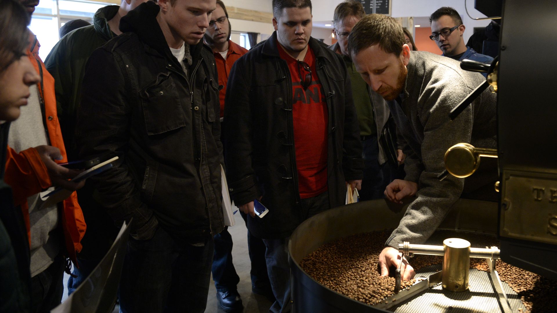 Group of men in jackets observe a coffee roasting machine as a staff member adjusts the roast; brown coffee beans roast in a large industrial setup.