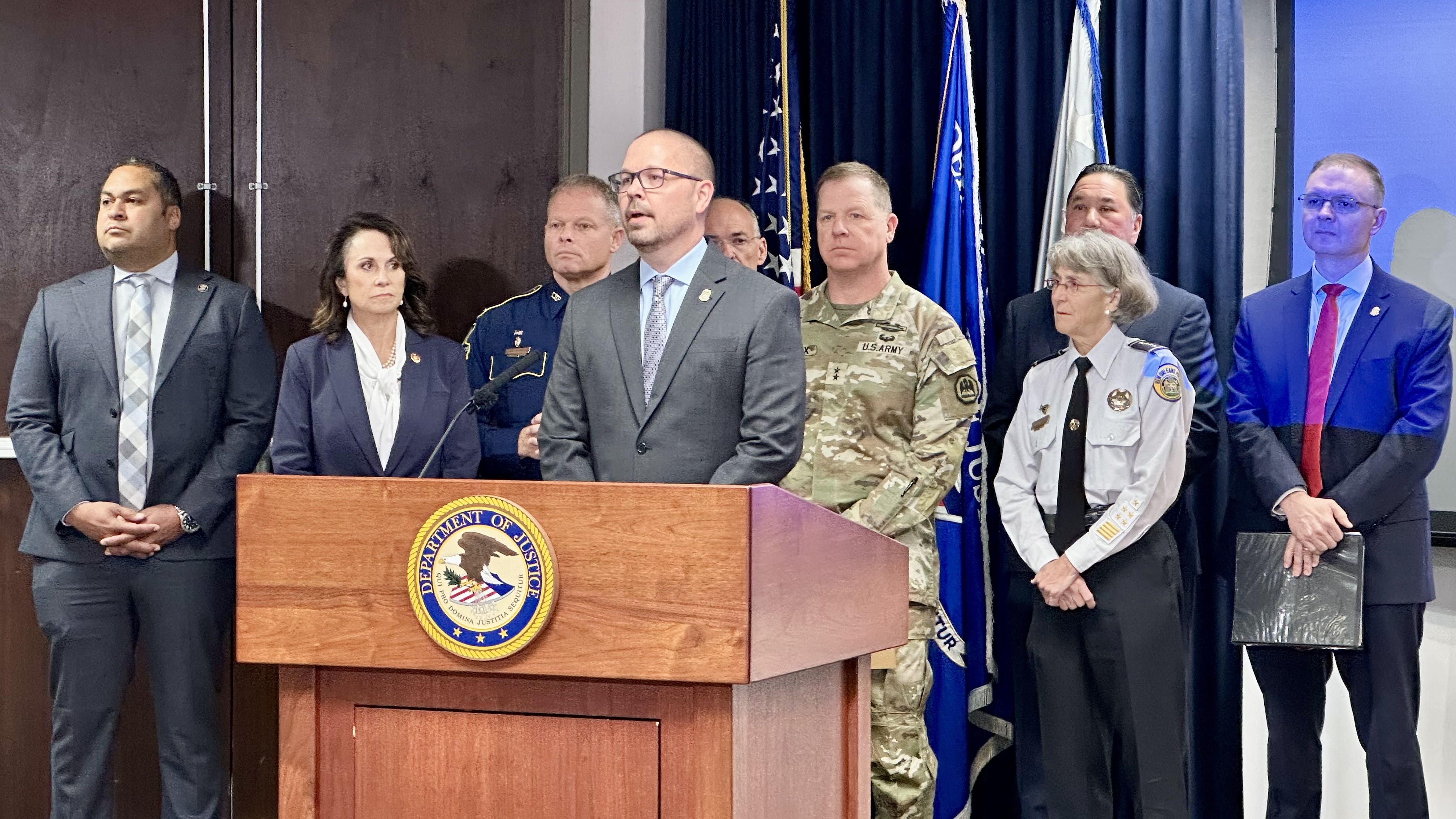 Group of officials and uniformed personnel stand behind a podium with the Department of Justice seal during a press conference with American flags in the background.