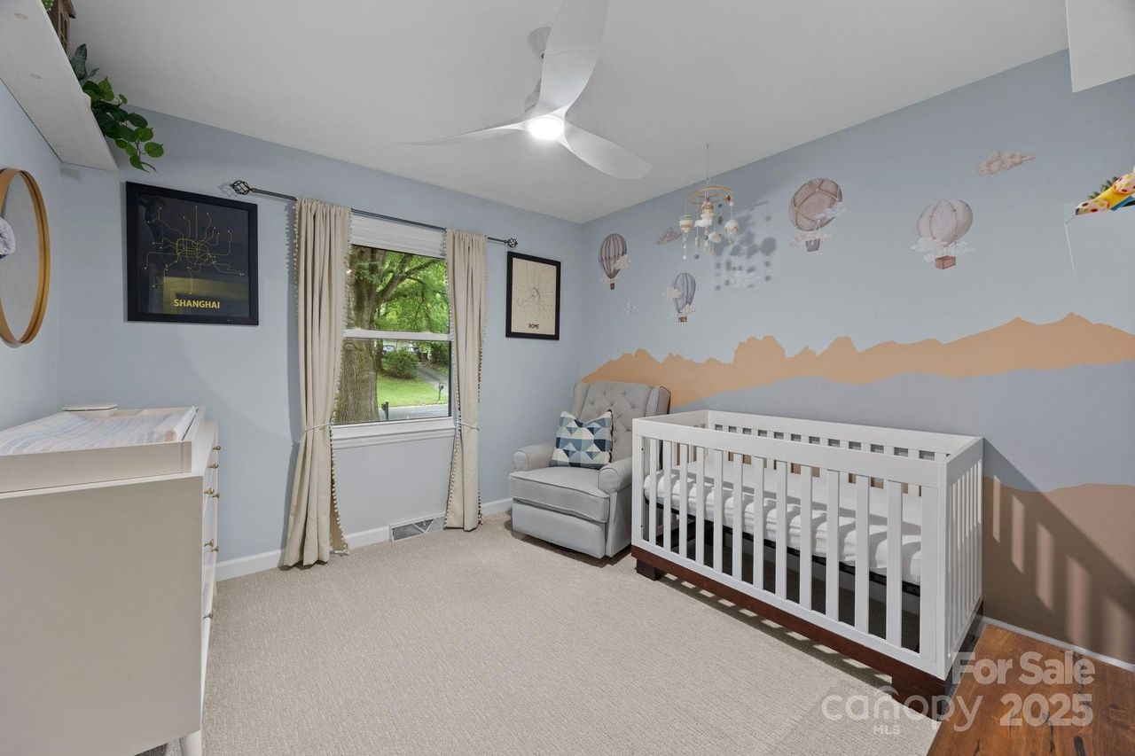 Light blue nursery with white crib, gray armchair with patterned pillow, beige curtains, and wall art of hot air balloons and mountain silhouette under ceiling fan.