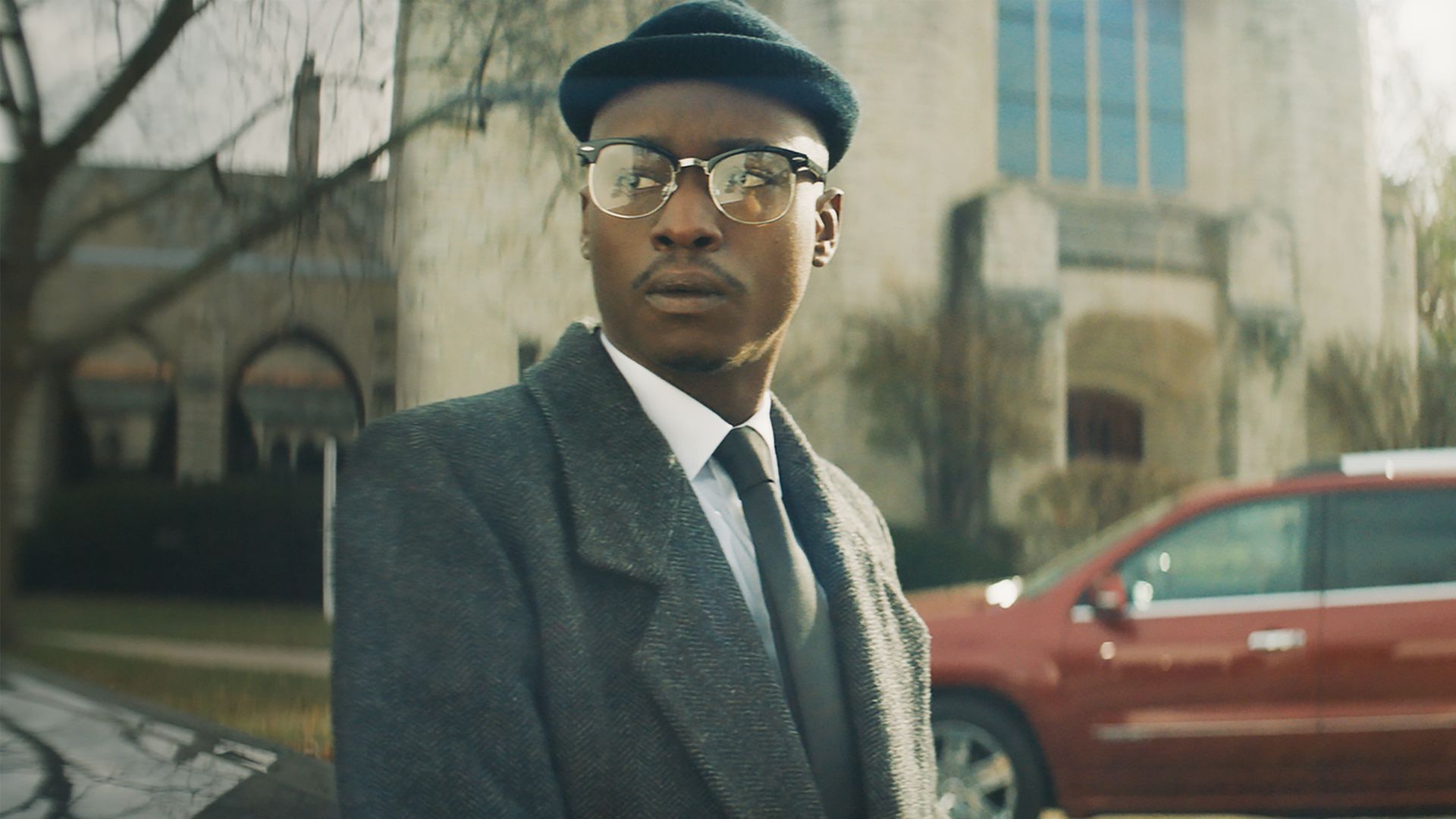 A stylish man wearing a dark tweed overcoat, white shirt, black tie, round glasses, and a navy cap stands outdoors by a red SUV and a stone building on a sunny day.