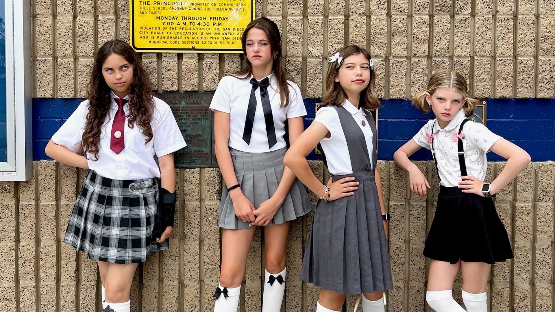Four young girls stand against a school looking annoyed and disaffected