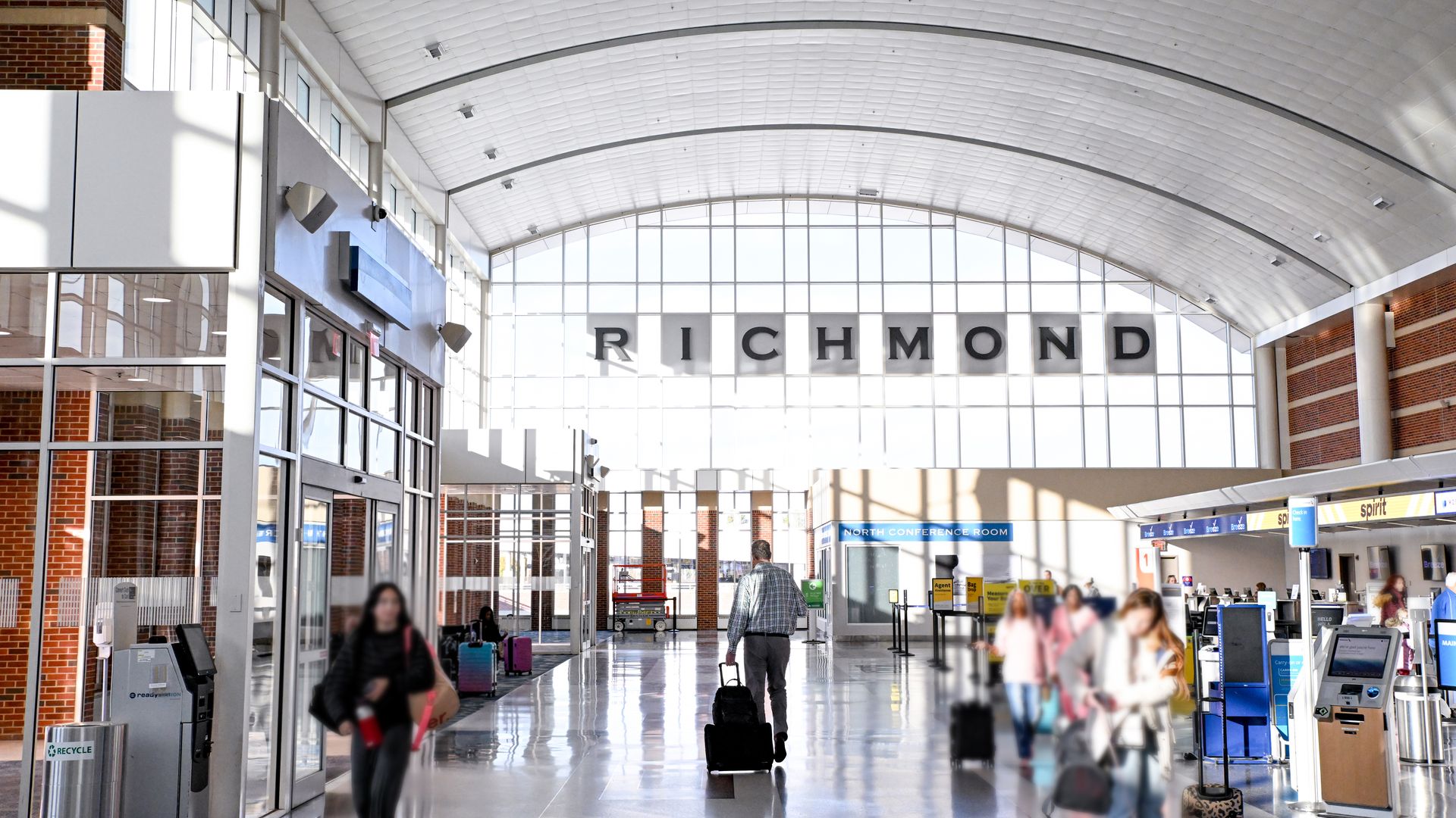 Bright airport concourse with a high curved white ceiling, glass walls and brick columns. A large Richmond sign stretches across the far end as travelers with luggage walk through.