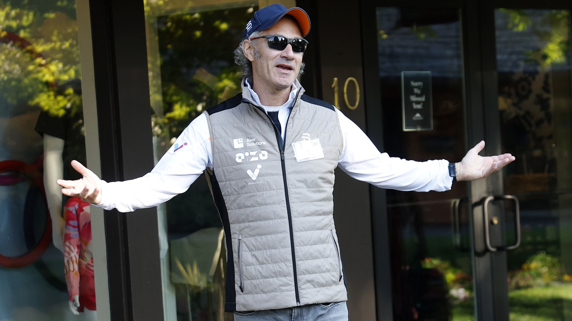 Man wearing sunglasses, a blue and orange cap, gray vest, and blue jeans, standing outdoors with arms outstretched in front of glass doors.