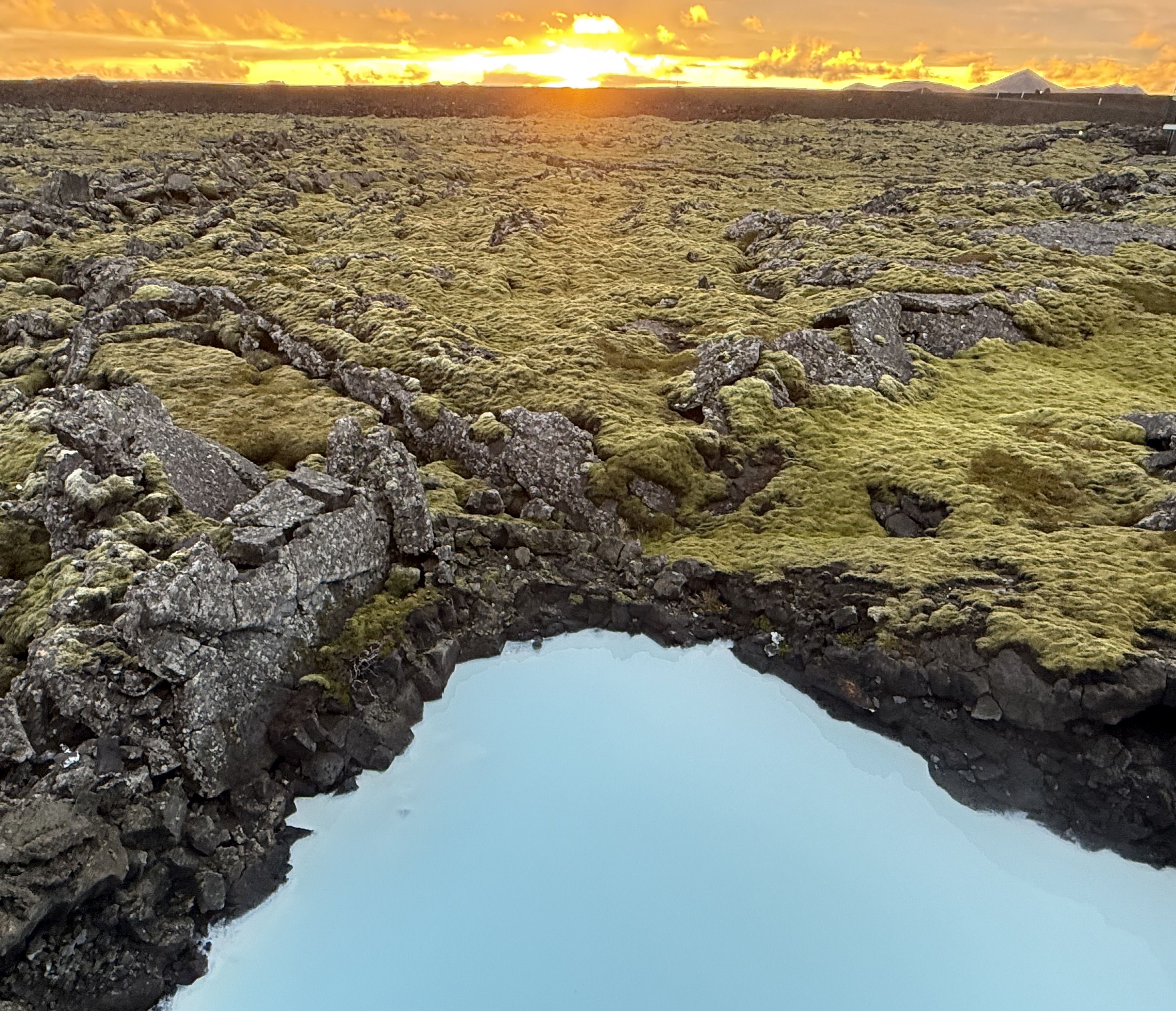 Sunset over a rugged lava field covered in green moss, with a dark ridge along the horizon and distant tents on the right; orange sky, glowing sun, and scattered clouds.
