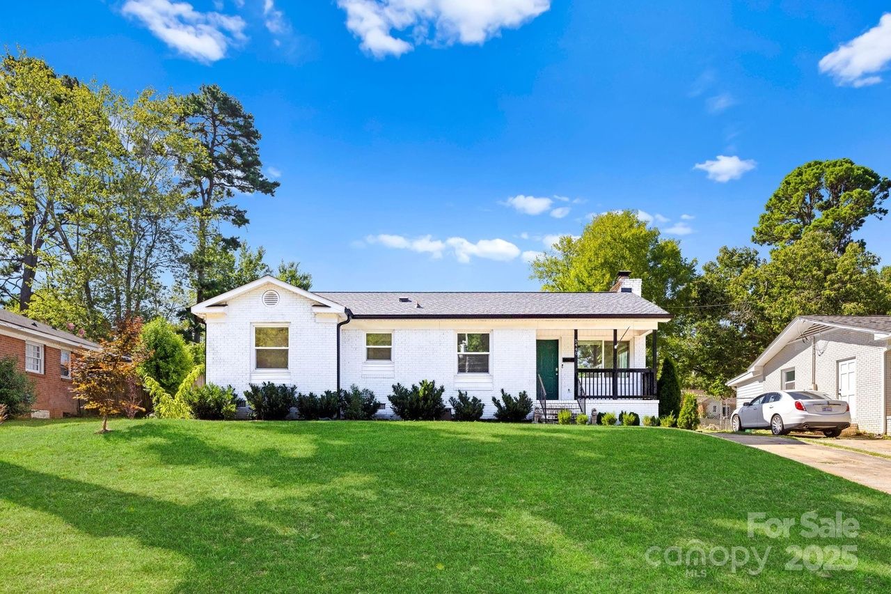 White brick single-story house with black trim and green door, large green lawn in front, blue sky with few clouds, trees surrounding property, and a white car parked on driveway.