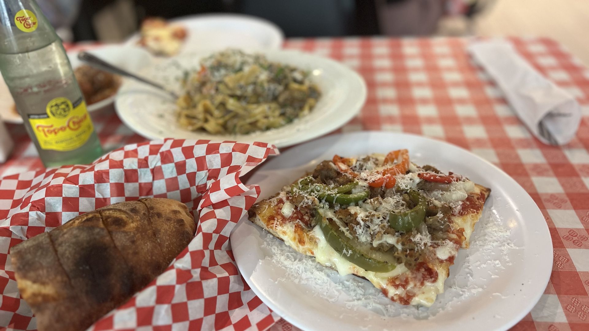 Table with a red-checkered cloth. Foreground shows a veggie pizza slice with peppers, onions, and mushrooms on a white plate; a crusty bread loaf sits in red checkered paper. Background shows pasta and a bottle of Topo Chico.