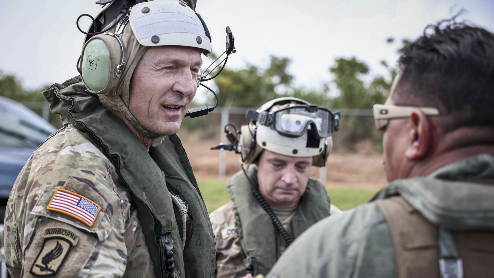 Gen. Randy George is clad in flight gear. He is smiling and standing two men. He touches one of their shoulders.