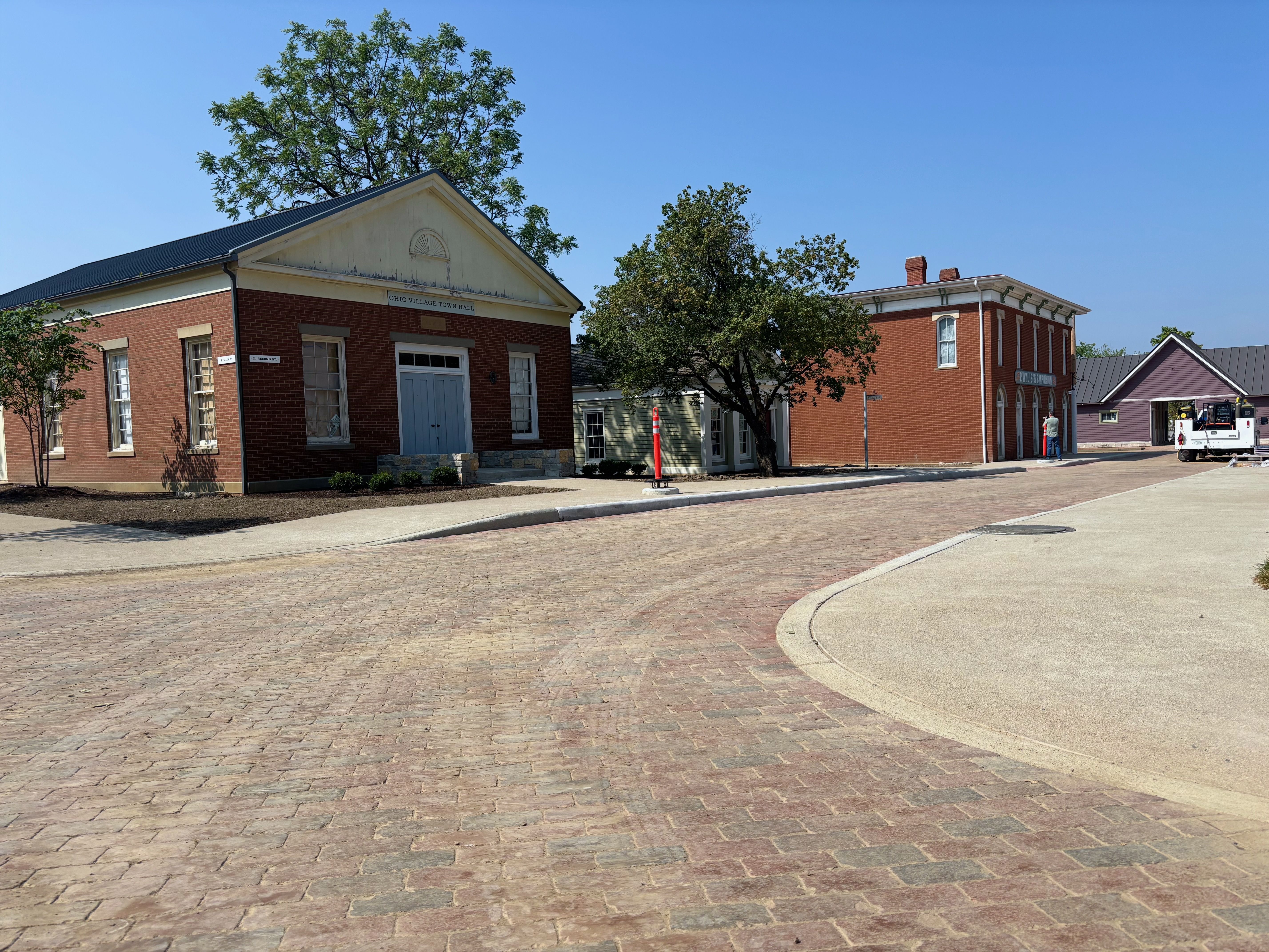 Bright sunny street with red brick town hall buildings, green trees, and a curved brick road. A pale pediment and blue door mark the town hall; a person by the right building.