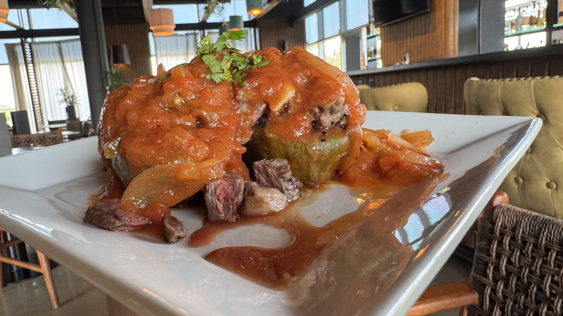 A stuffed, fried avocado topped with red tomato sauce and garnished with herbs is served on a square white plate with chunks of beef and onions. The dish is photographed in a modern restaurant setting.