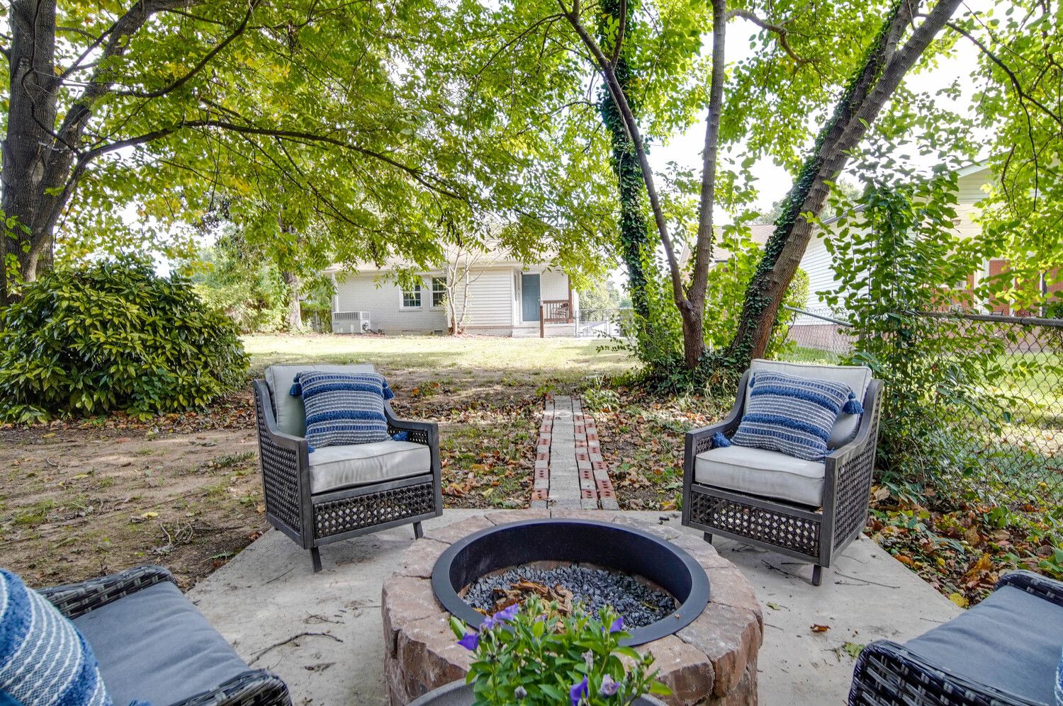 Cozy backyard patio under a leafy canopy with four gray wicker chairs and blue striped pillows arranged around a circular stone fire pit. A brick path leads to a house in the background.