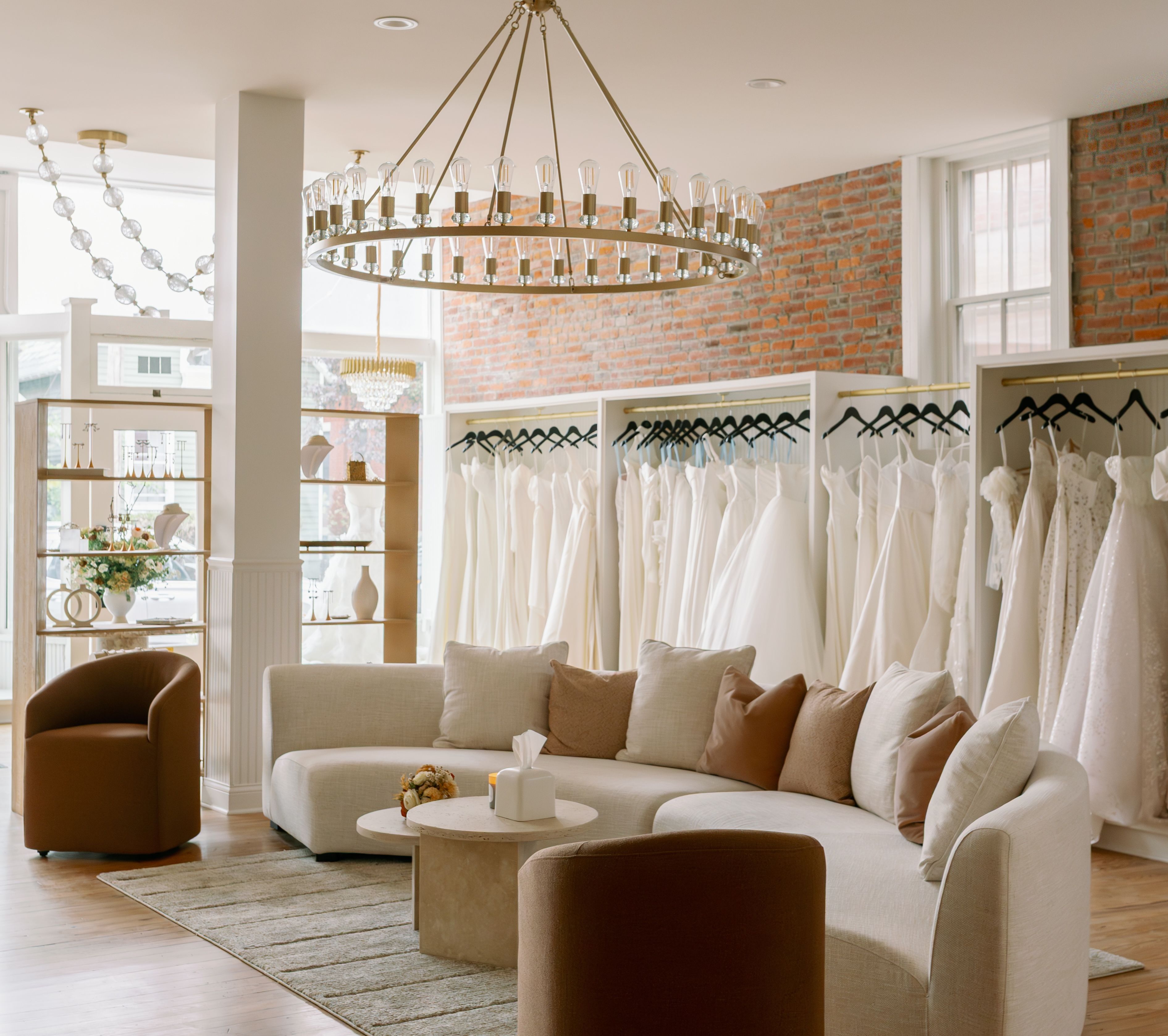 Bright bridal boutique interior with white wedding dresses hanging on racks, exposed brick wall, beige curved sofa with pillows, brown armchairs, and a large chandelier overhead.