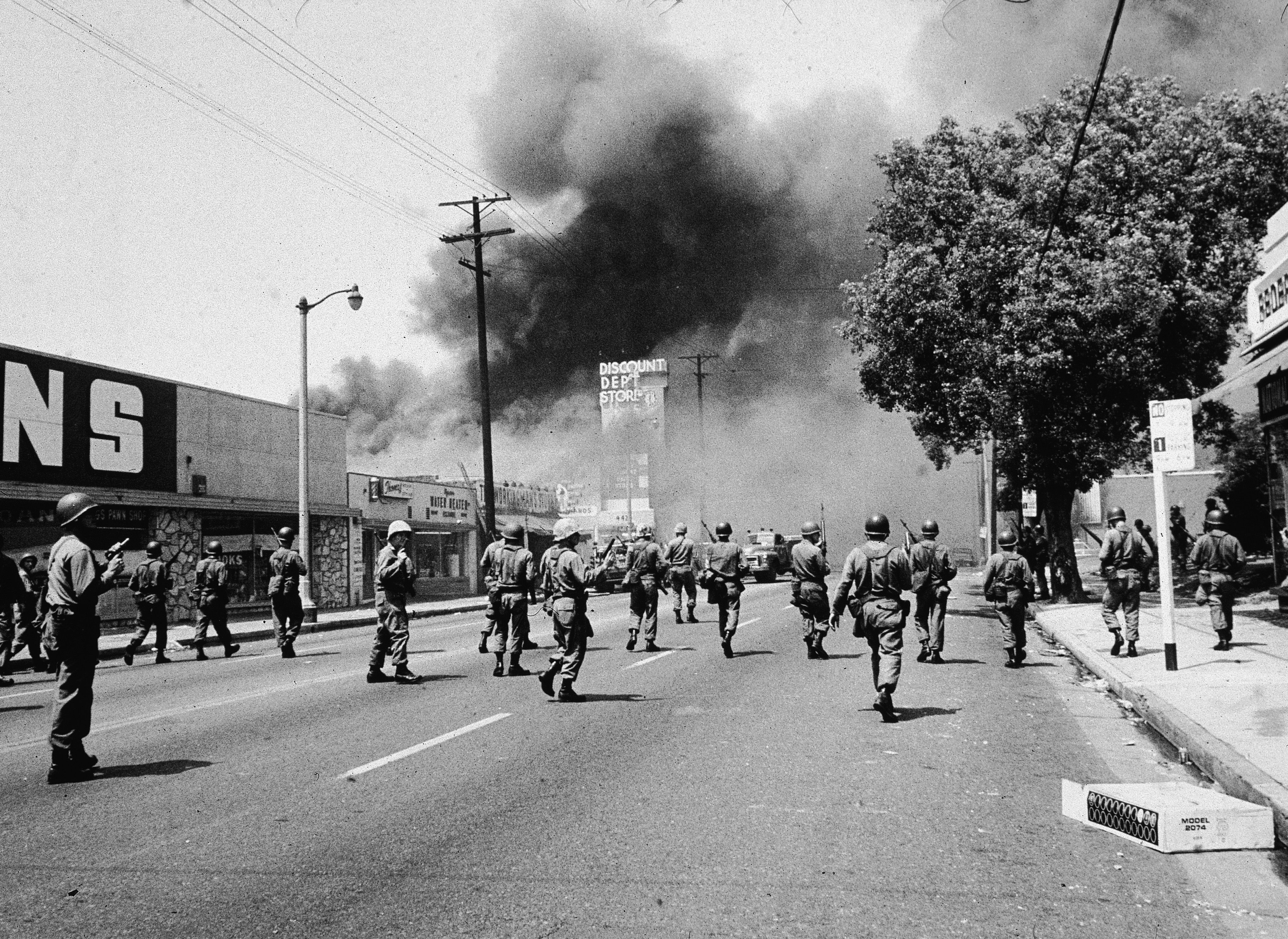 Black-and-white photo of armed soldiers walking on a street with heavy smoke rising from a fire near a discount department store and other businesses.
