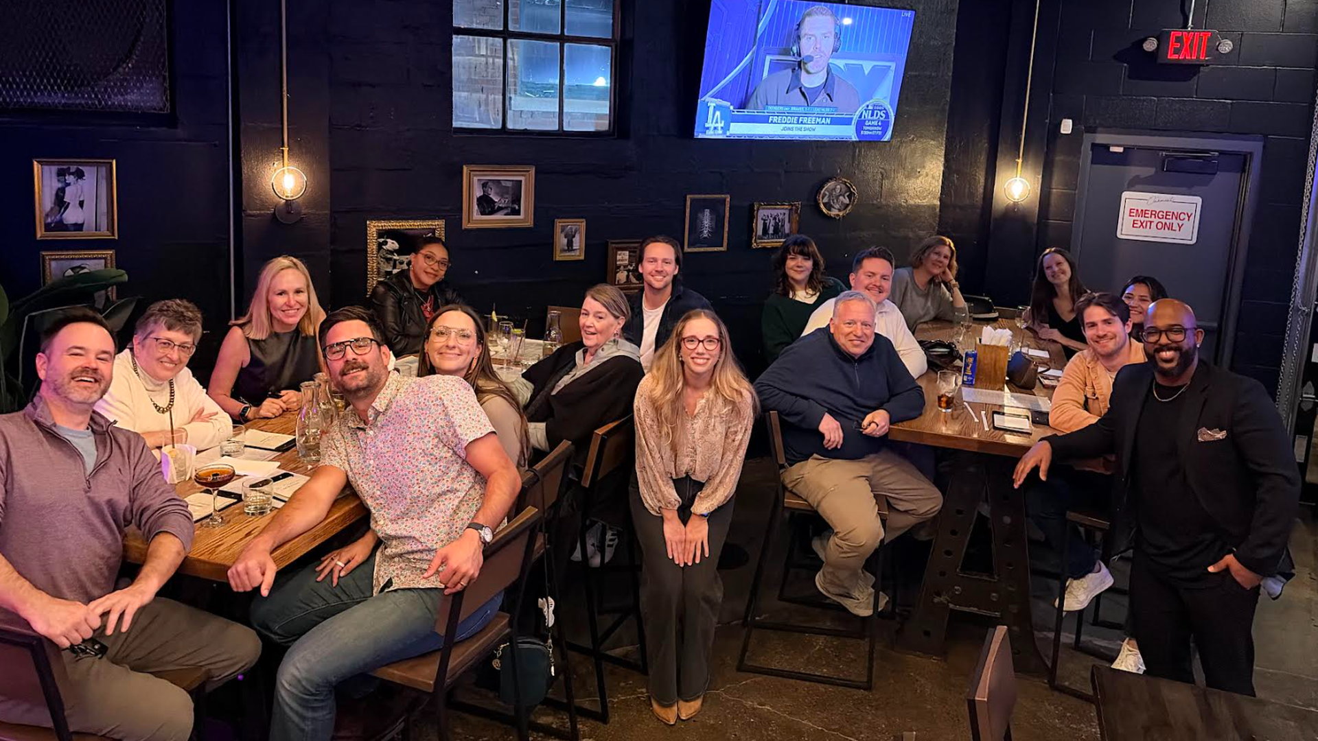 Group of about 15 people around a long wooden table in a dim bar, smiling at the camera. A wall TV with a sports broadcast, framed photos on dark walls, and an emergency exit sign is visible.