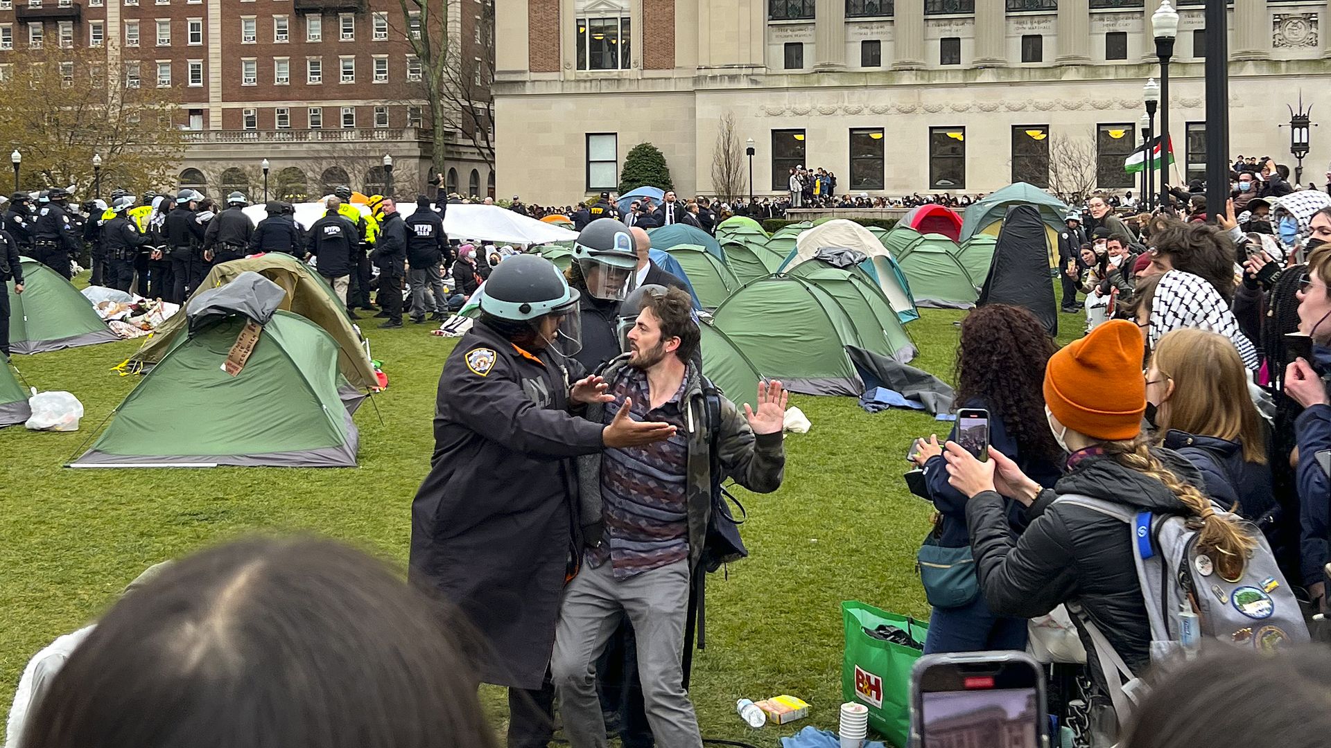 Person is being detained by police officers in riot gear. To the right, dozens of people are looking. In the background, a protest encampment is seen with green tents. More officers are in the back of the photo.