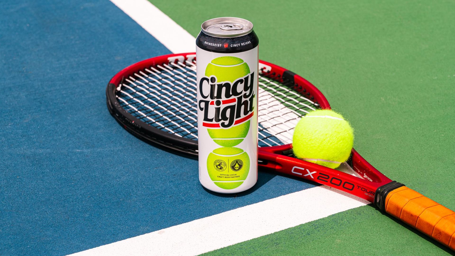 A can of "Cincy Light" beer sits upright on a red tennis racket on a blue and green tennis court, with a bright yellow tennis ball nearby.