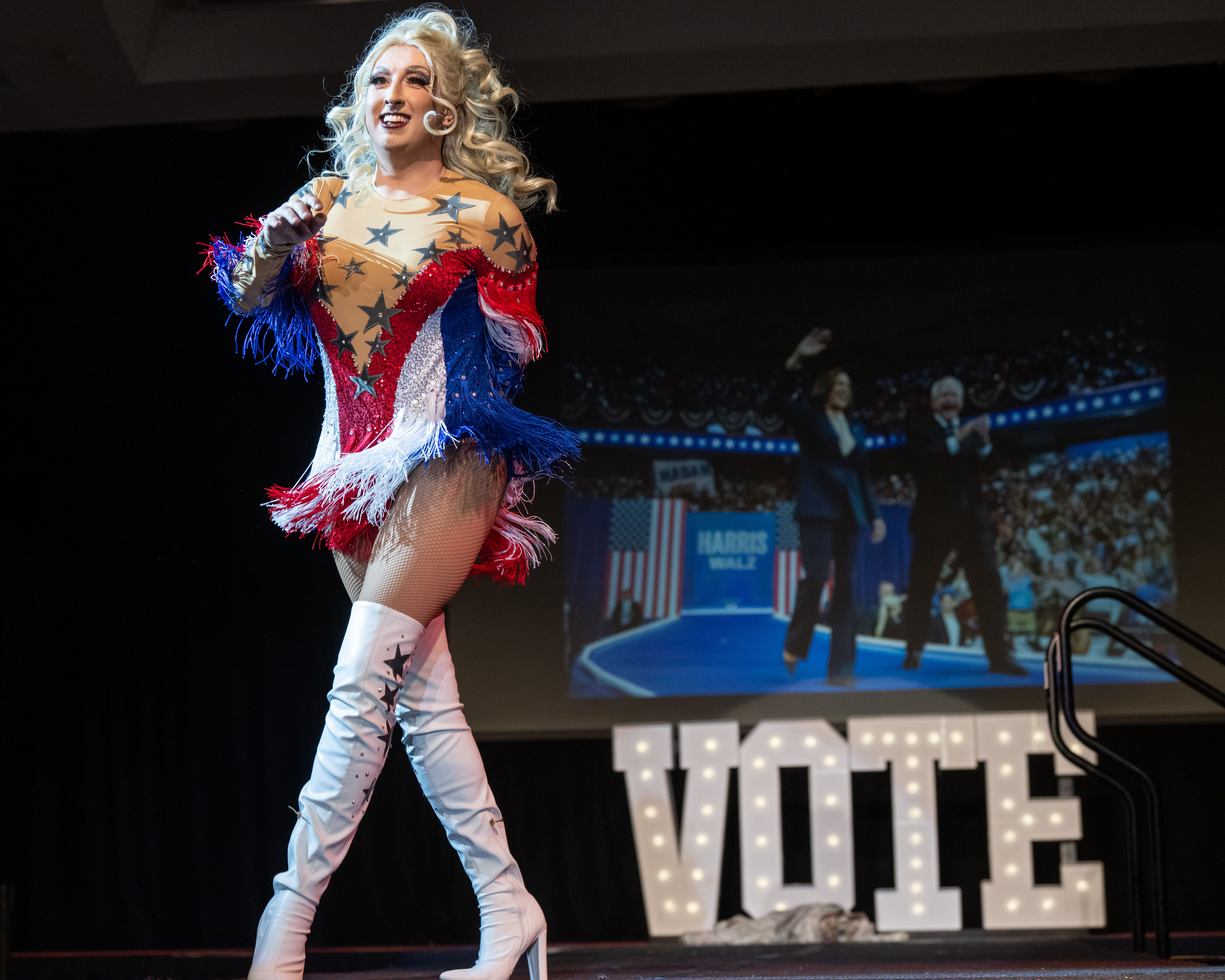 A drag performer in a star-spangled red, white and blue costume with fringe and blonde curls smiles on stage, raising a fist as a cheering audience watches from the background.
