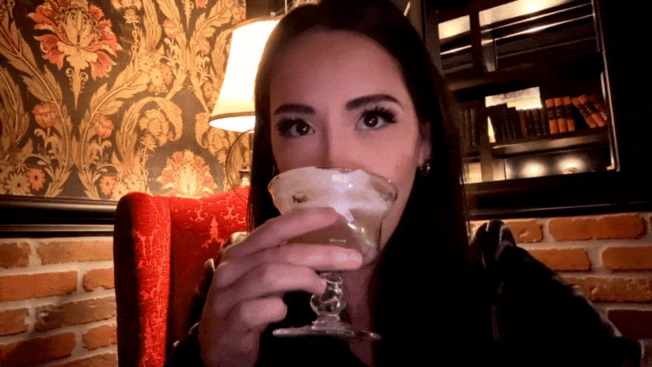 Woman with dark hair drinks from a decorative glass in a cozy room with floral wallpaper, a red cushioned chair, a lamp, and brick wall with bookshelves in the background.