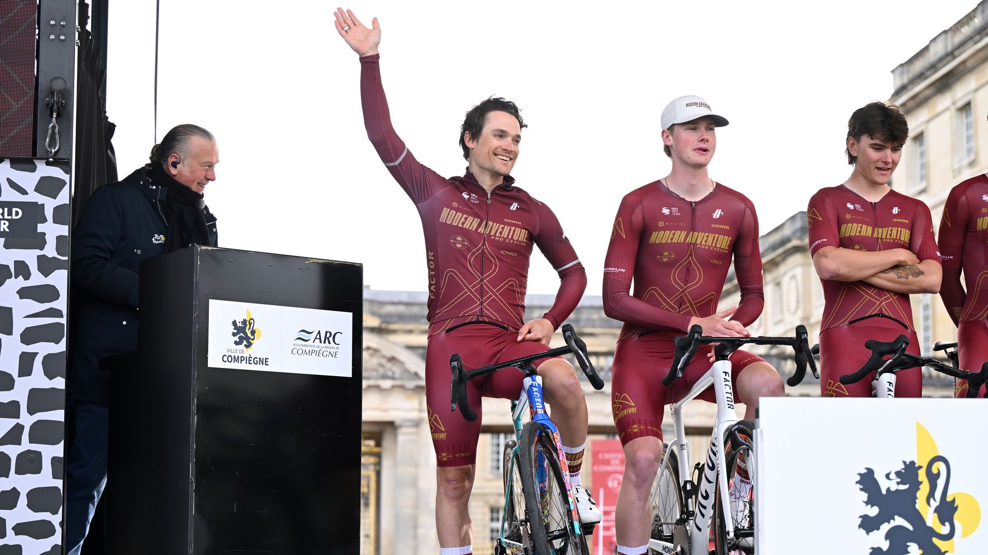 Three cyclists in burgundy kits stand with their bikes on a podium; the center rider waves as a man at a nearby podium smiles, with historic buildings in a city square behind them.