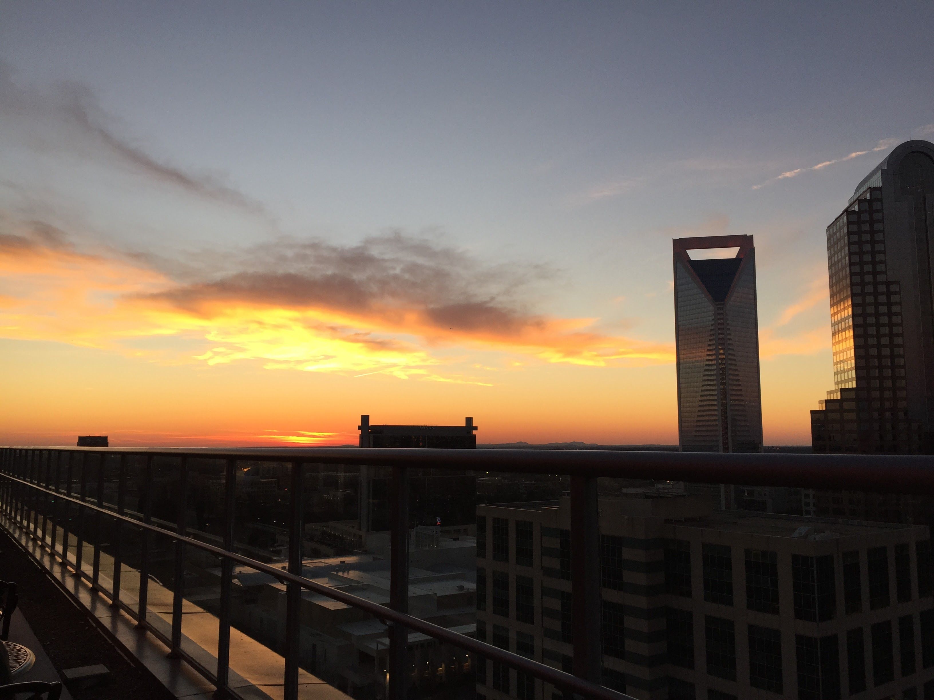 City skyline at sunset with orange and yellow clouds, a tall building with triangular top, and glass railing in foreground on rooftop.