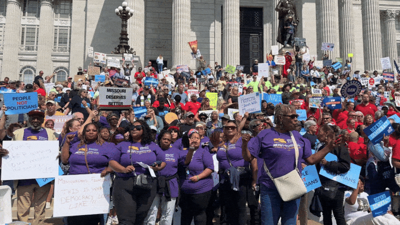 A large group of protesters on government building steps holding signs like "PEOPLE NOT POLITICIANS," "MISSOURI DESERVES BETTER," many wearing purple shirts and red shirts.