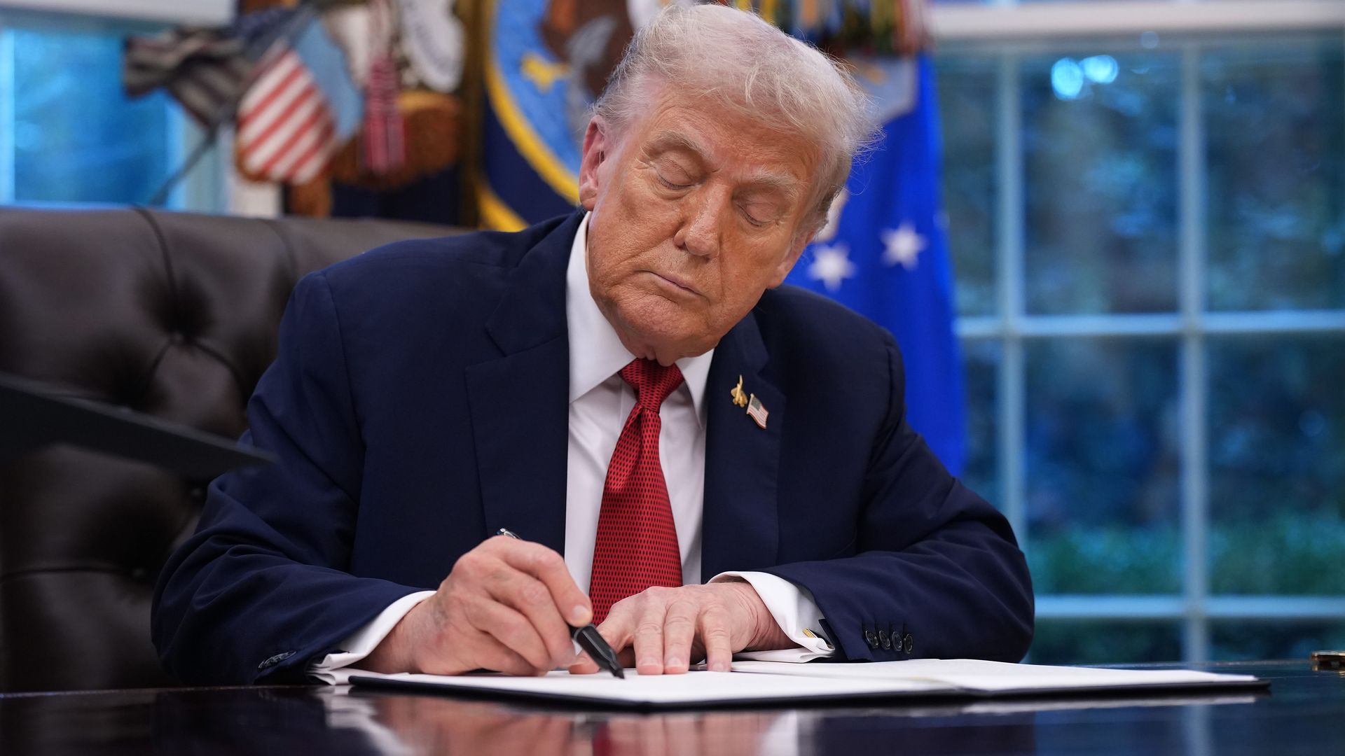 President Donald Trump sits at a desk in the Oval Office signing an executive order, with papers and a pen in front of him.