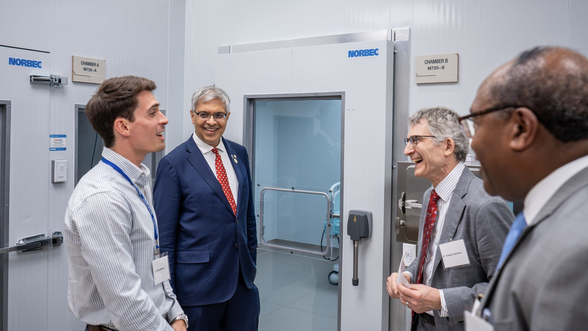 Four professionals in business attire with badges stand in a clean, metallic lab, near glass-front walk-in chambers labeled NORBEC and Chamber A/B, smiling and chatting.