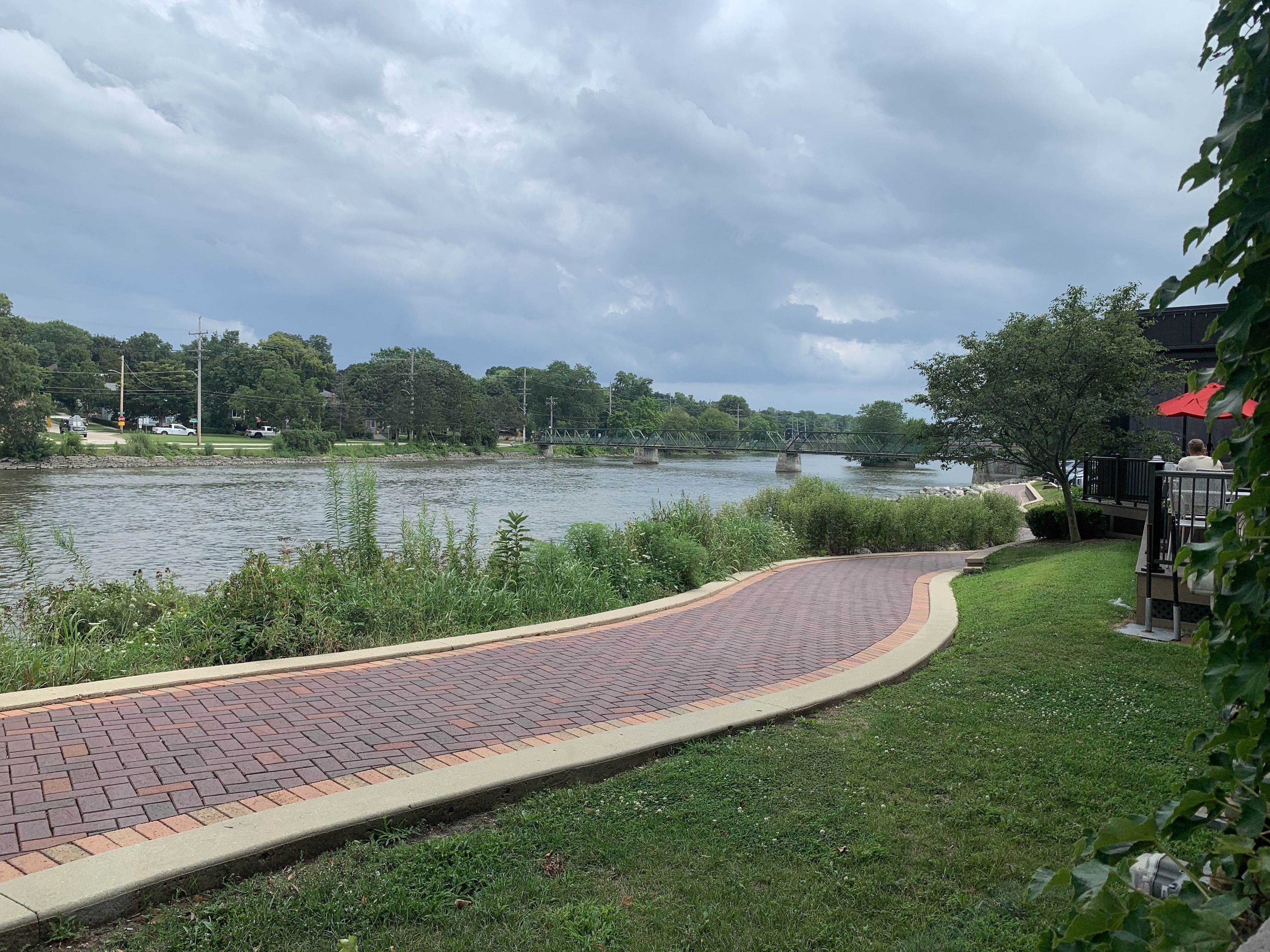 Riverside park with a curved brick walkway along the water, green trees, and a distant metal footbridge. Dark storm clouds fill the sky; red umbrellas mark a seating area on the right.
