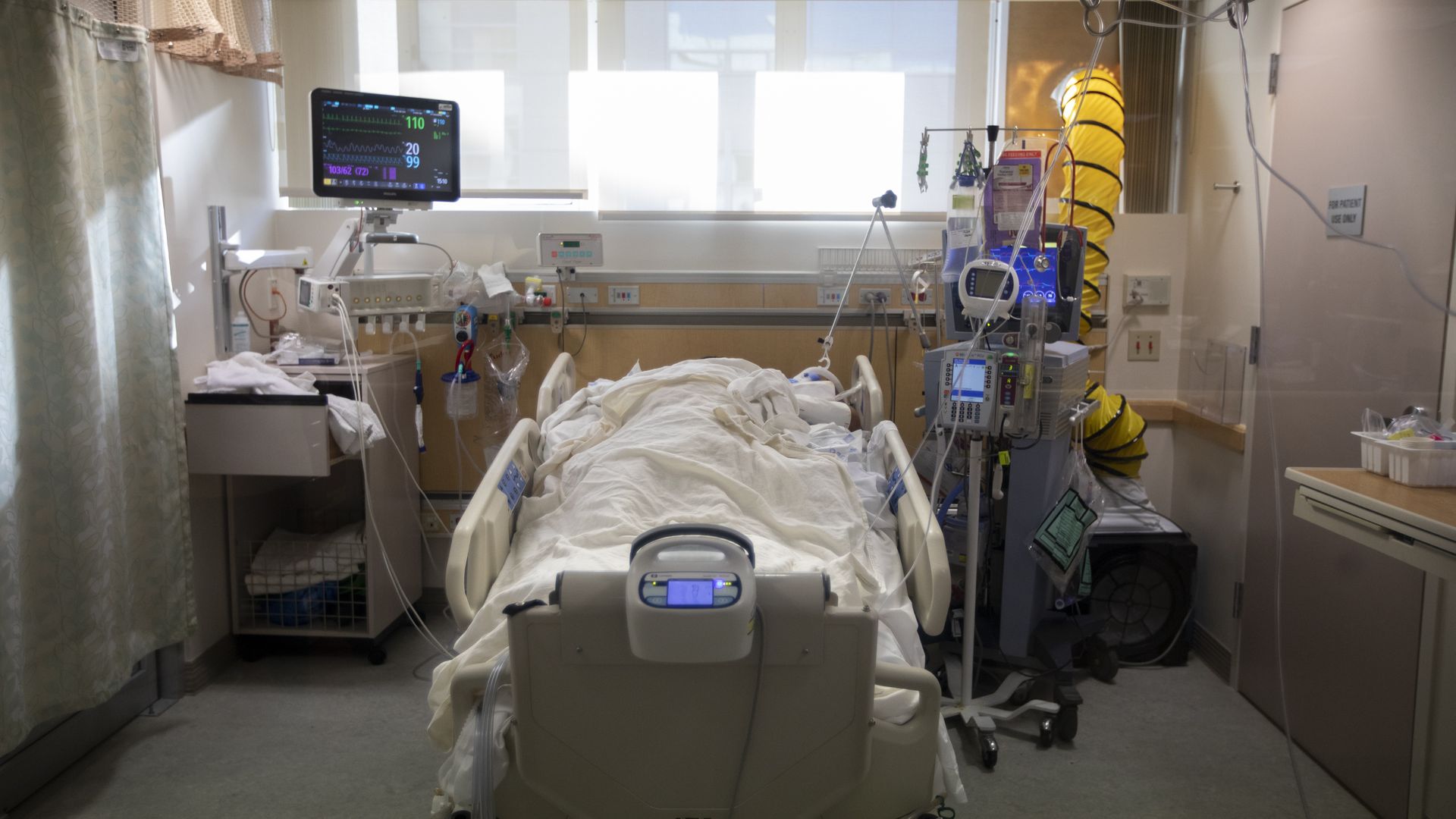 A patient lays fully covered in a white hospital bed with monitors on each side.