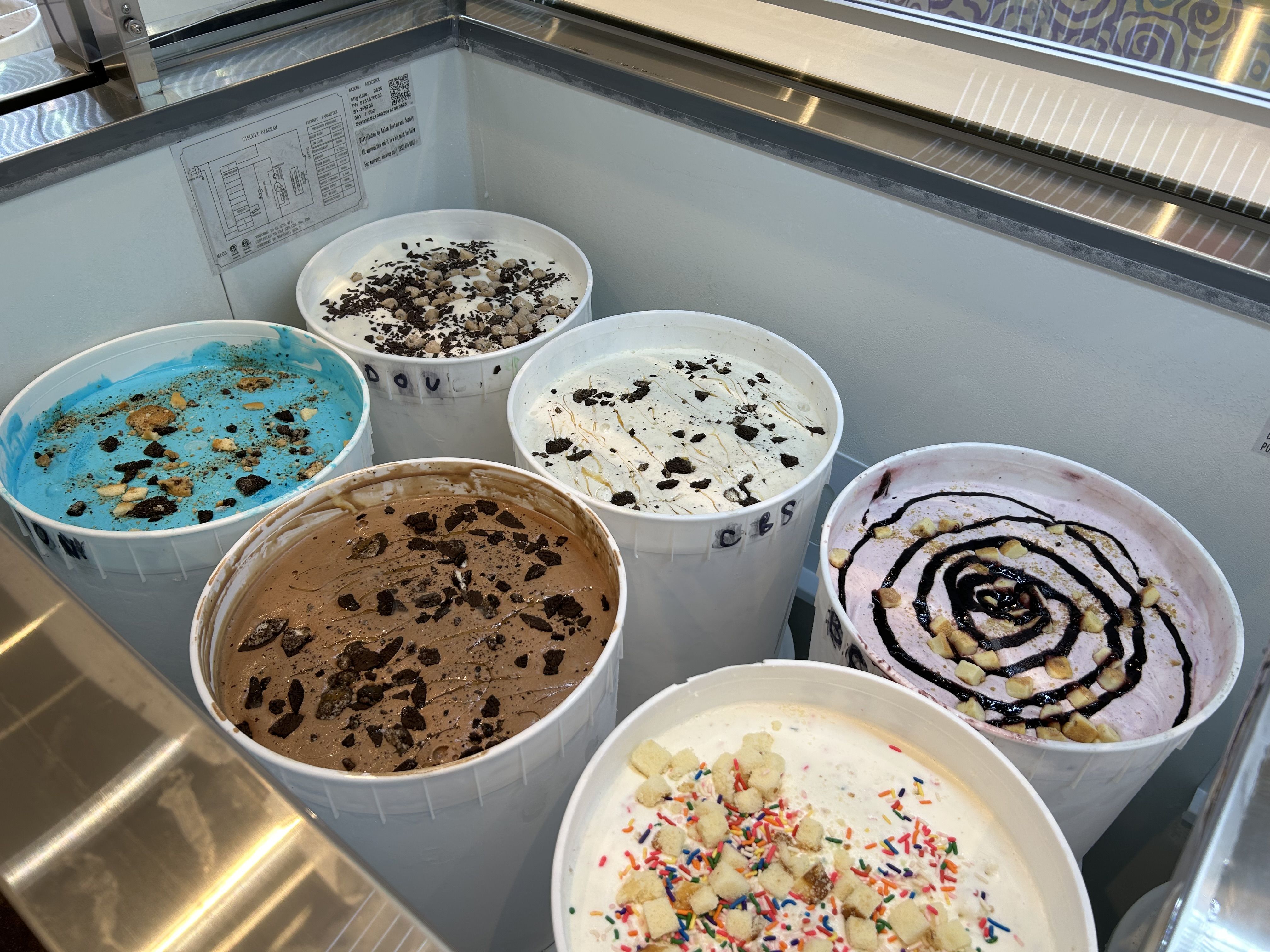 Six large white tubs of colorful ice cream with various toppings like cookie pieces, chocolate drizzle, and sprinkles, displayed in a freezer at an ice cream shop.
