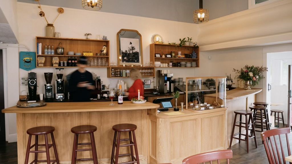 A warm, light-filled cafe interior with wooden counters and bar stools, espresso machines, pastries under glass, and floral arrangements. Two staff members serve drinks behind the bar.