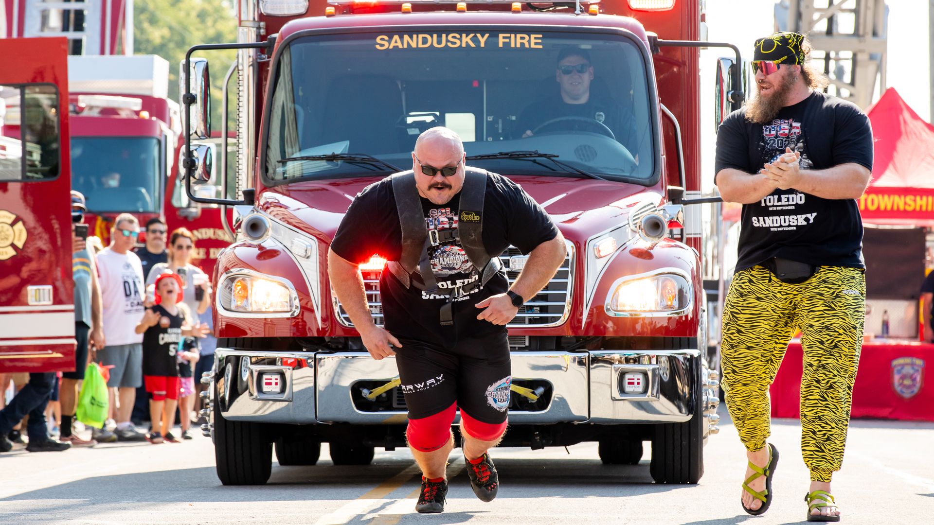 A man wearing pulls a Sandusky fire truck using a harness while another man claps beside him during an outdoor event.