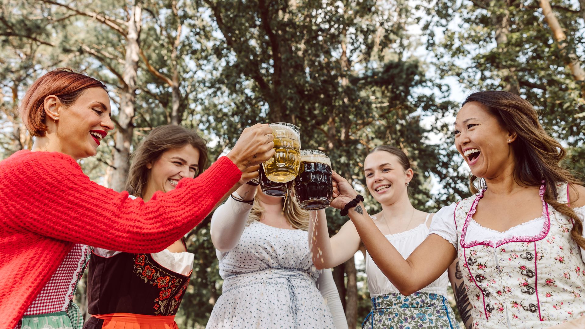 Group of women outdoors in a sunny forest, toasting beer mugs. They wear colorful outfits, including a bright red sweater and floral dirndl-style dresses, smiling and cheering.