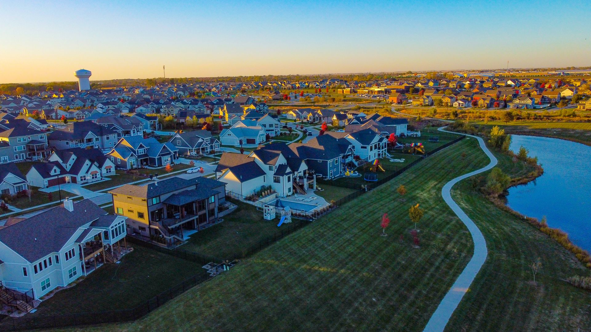 An aerial view of a sprawling suburban neighborhood at golden hour: rows of blue-gray houses, winding roads, a water tower in the distance, and a curving path along a lake with autumn trees.